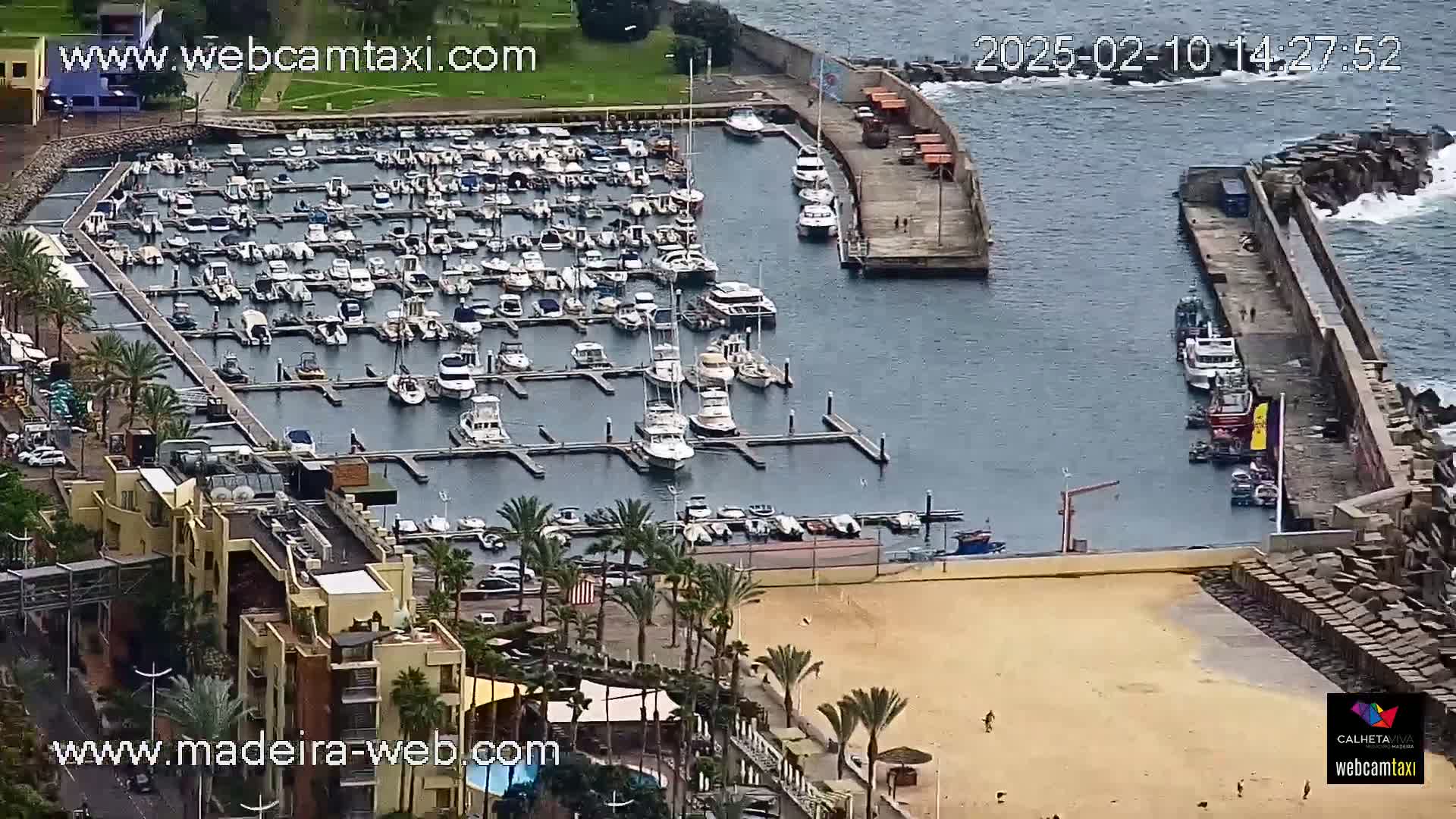 An aerial view shows a marina filled with numerous boats, adjacent to a sandy beach and buildings, on an overcast day.