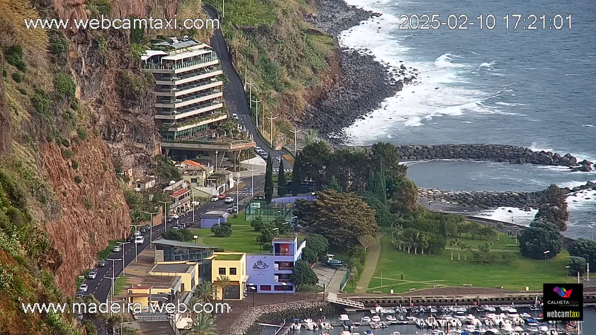 A coastal town scene shows a multi-story building on a cliff overlooking a small harbor filled with boats, a grassy park, and a rocky shoreline with moderate waves.