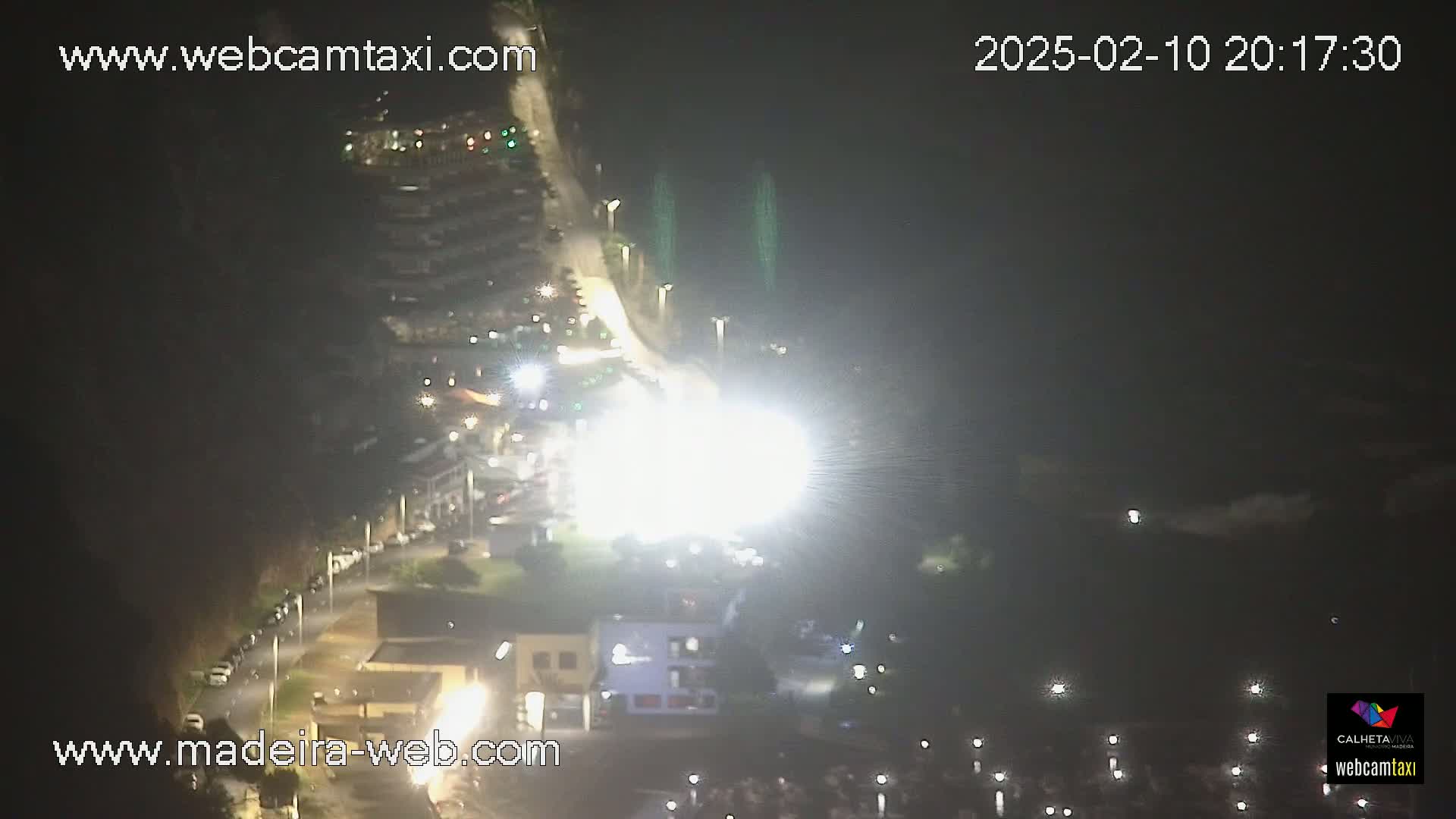 A nighttime aerial view shows a brightly lit area with cars parked along a road, buildings, and a body of water with lights reflecting on it.