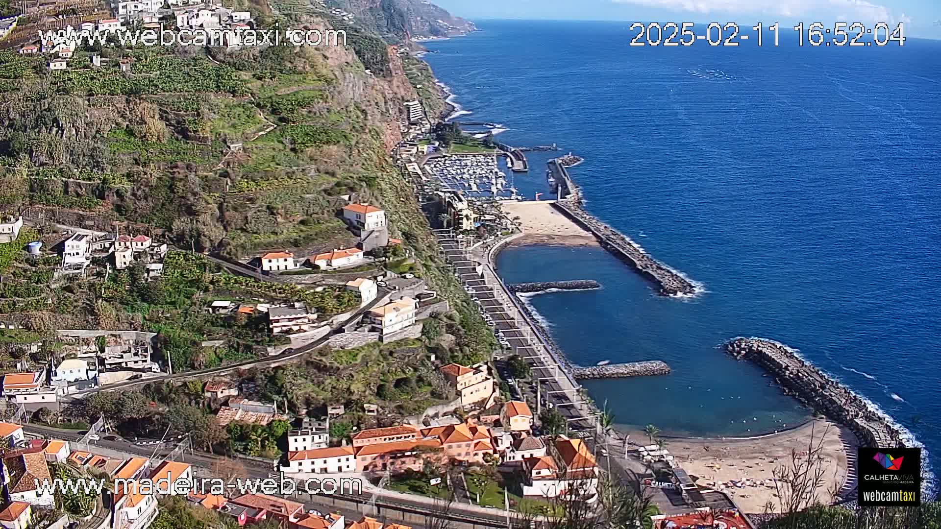 A coastal town with terracotta-roofed buildings climbs a steep hillside overlooking a calm ocean bay with a small marina and sandy beach under sunny skies.