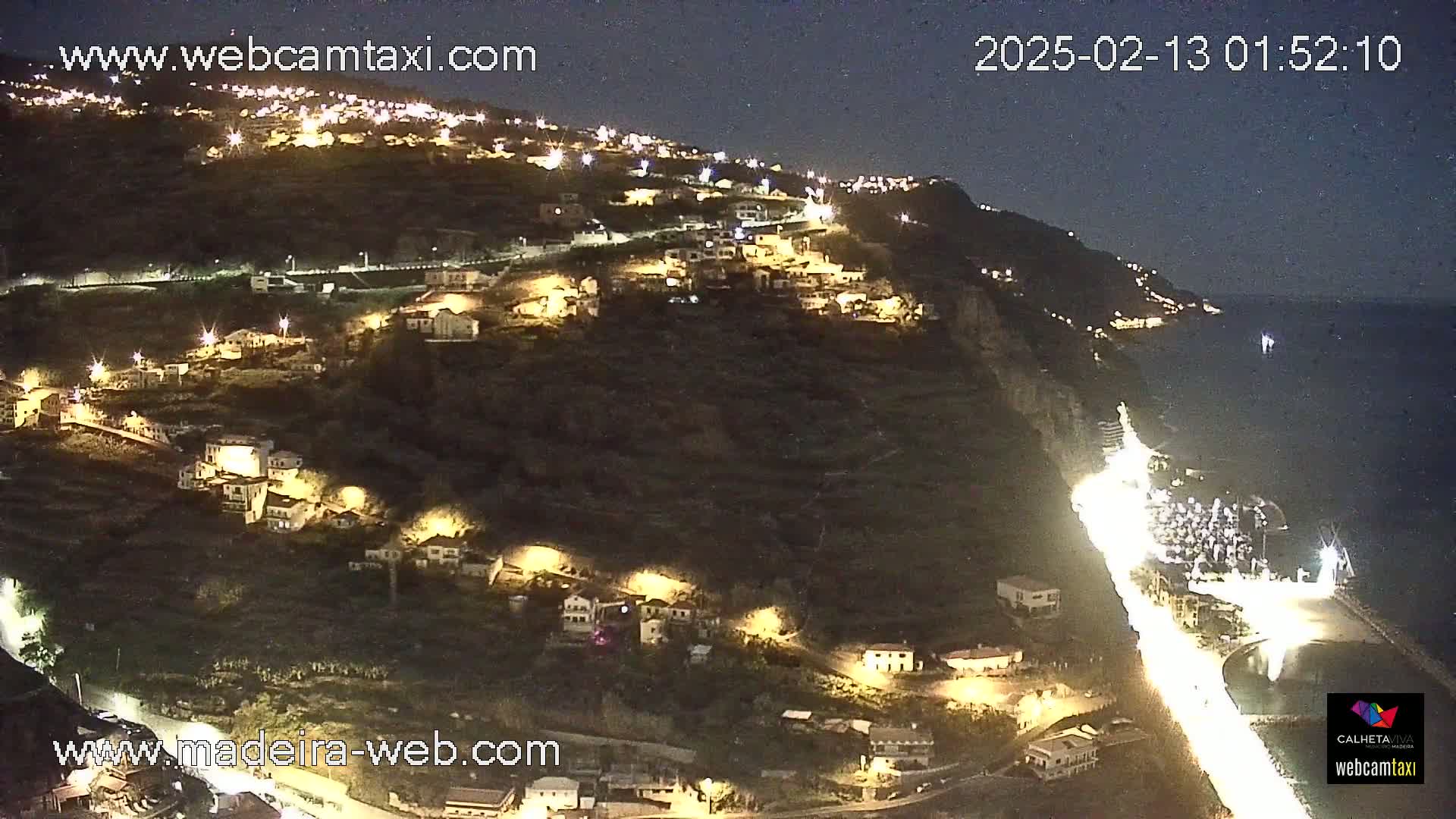 A nighttime view of a coastal town built on a hillside overlooking the ocean, with lights illuminating the buildings and roads.