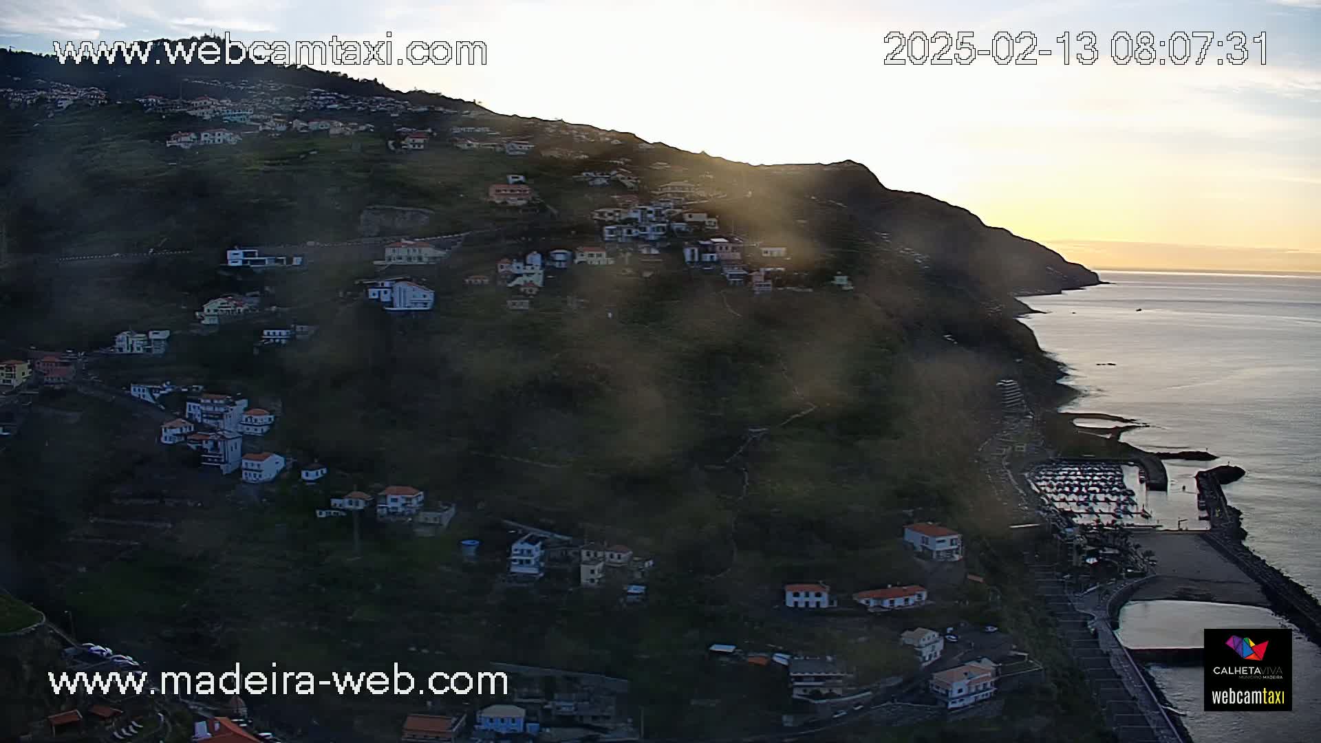 A coastal village nestled on a hillside overlooks a calm ocean at sunrise, with a light haze hanging in the air.