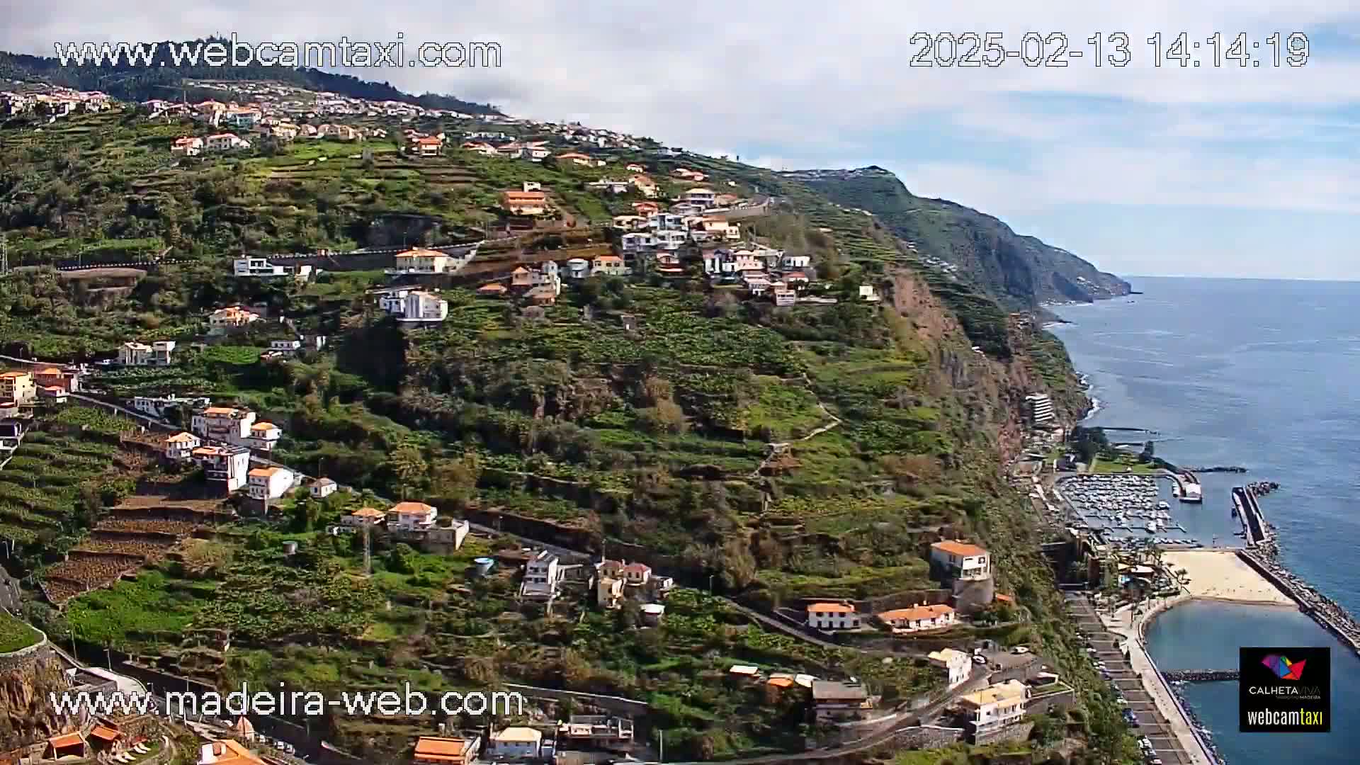 A hillside village with terraced farmland overlooks a calm ocean and a small marina under a partly cloudy sky.