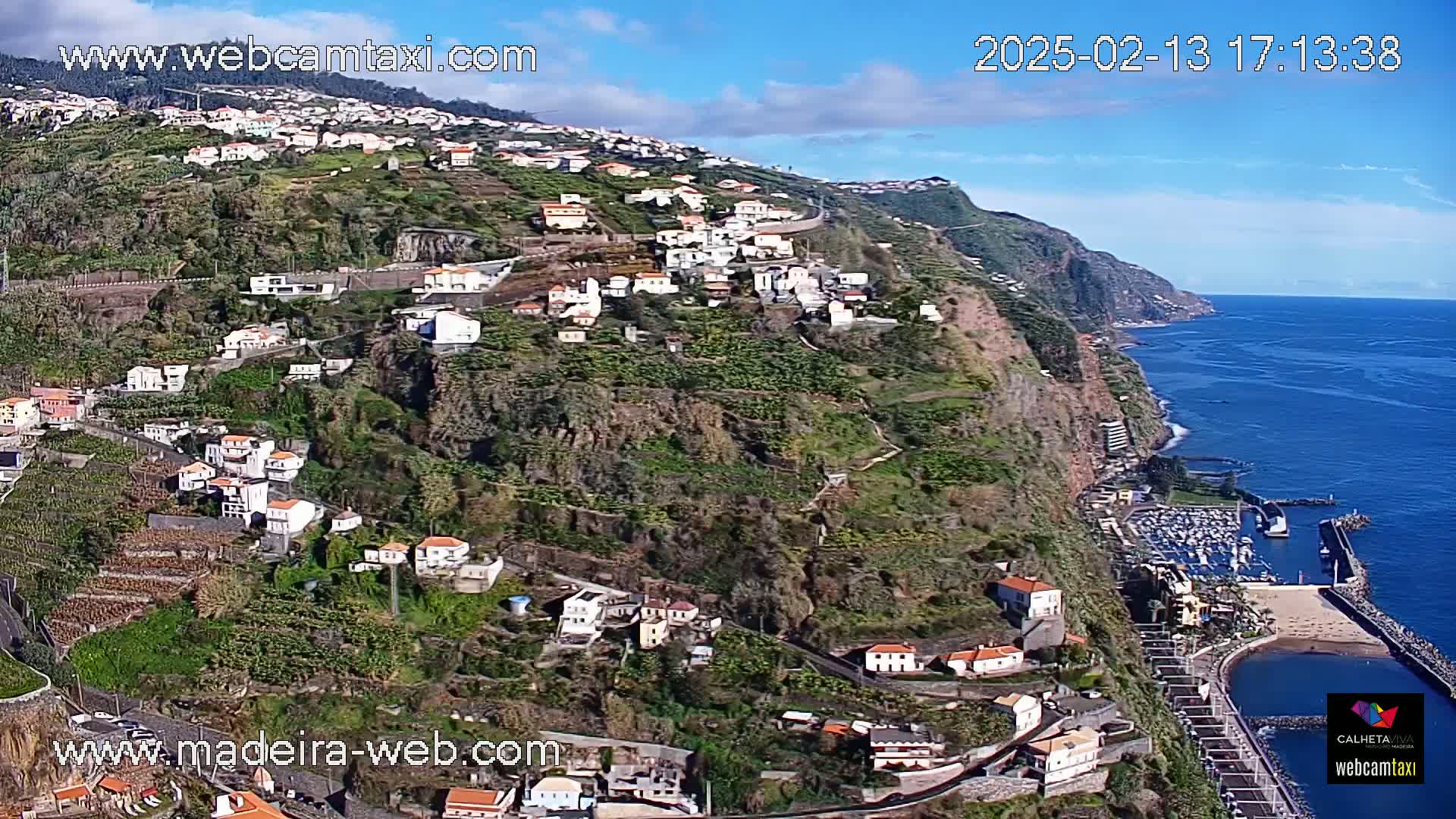 A coastal town clings to a steep, verdant hillside overlooking a calm ocean under a mostly sunny sky.