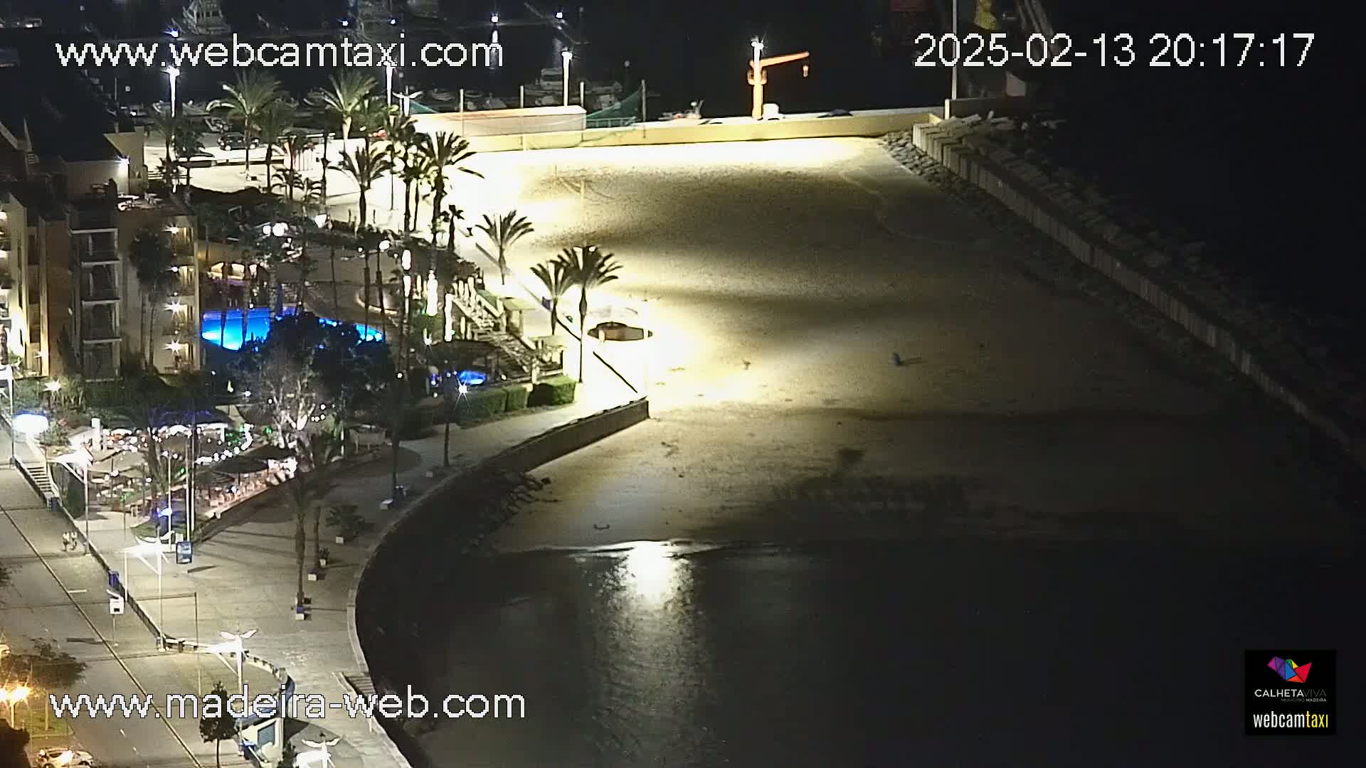 A nighttime aerial view of a beach illuminated by artificial lights, adjacent to a building with a swimming pool and palm trees, and calm dark water visible beyond the sand.