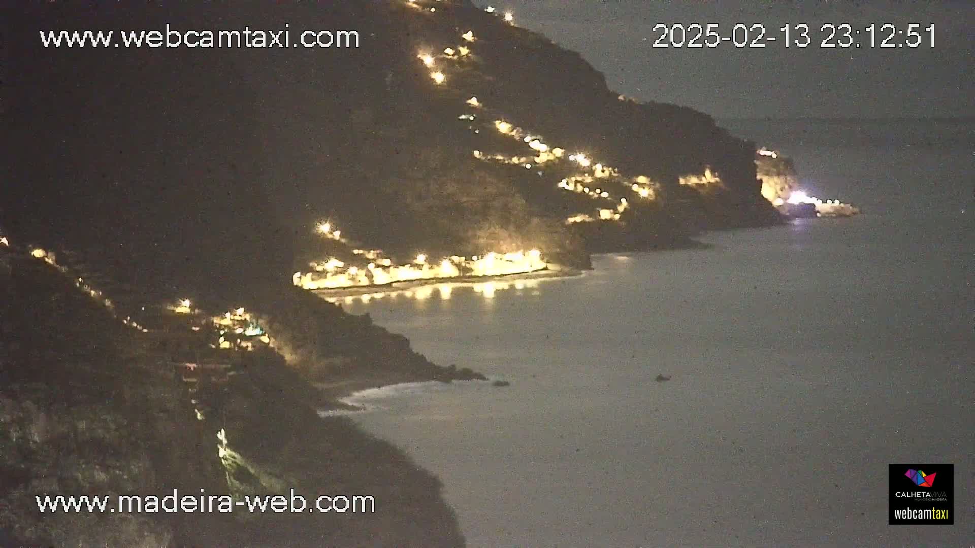 A nighttime, coastal scene shows a hillside town illuminated with lights along a dark body of water.