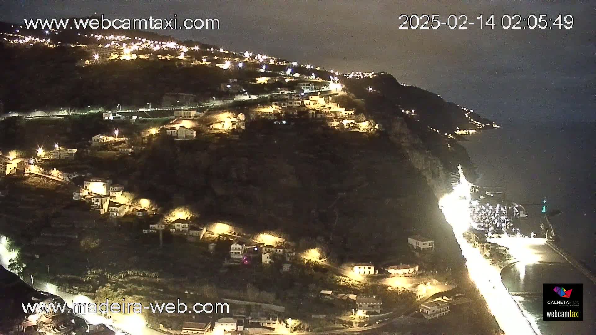 A nighttime, aerial view of a coastal town built on a hillside, with lights illuminating buildings and roads along the water.