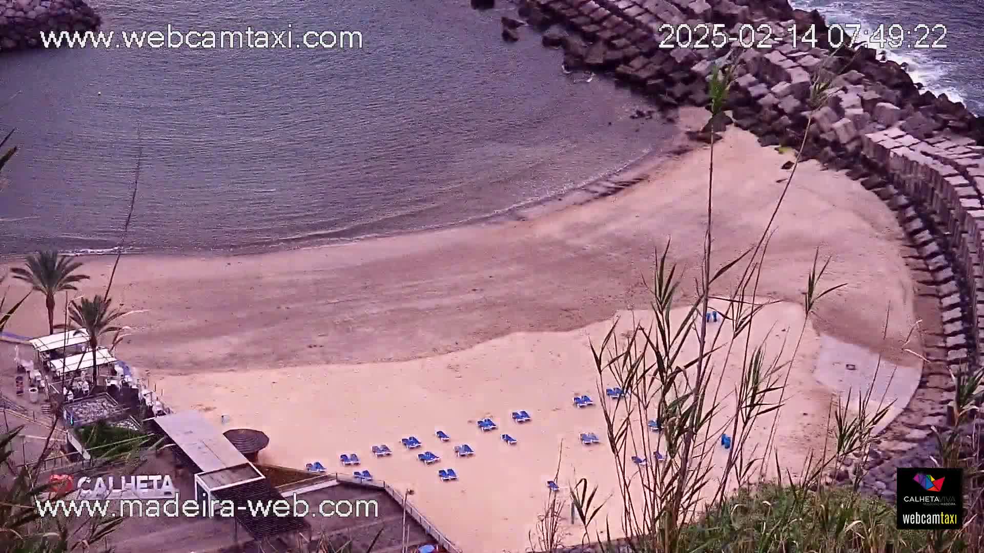 An overhead view of a sandy beach with several empty lounge chairs near calm ocean water, partially enclosed by a rock wall.
