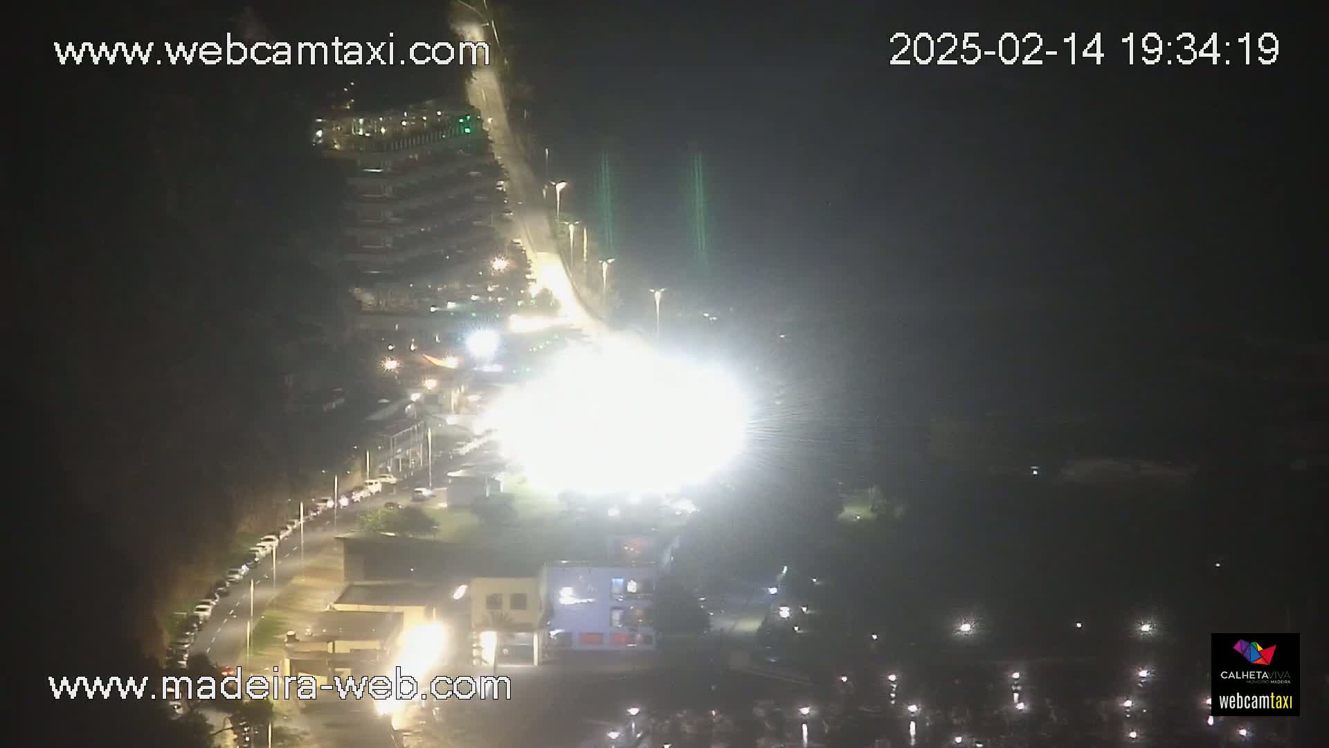 A nighttime aerial view of a brightly lit road with parked cars and buildings, under clear skies.
