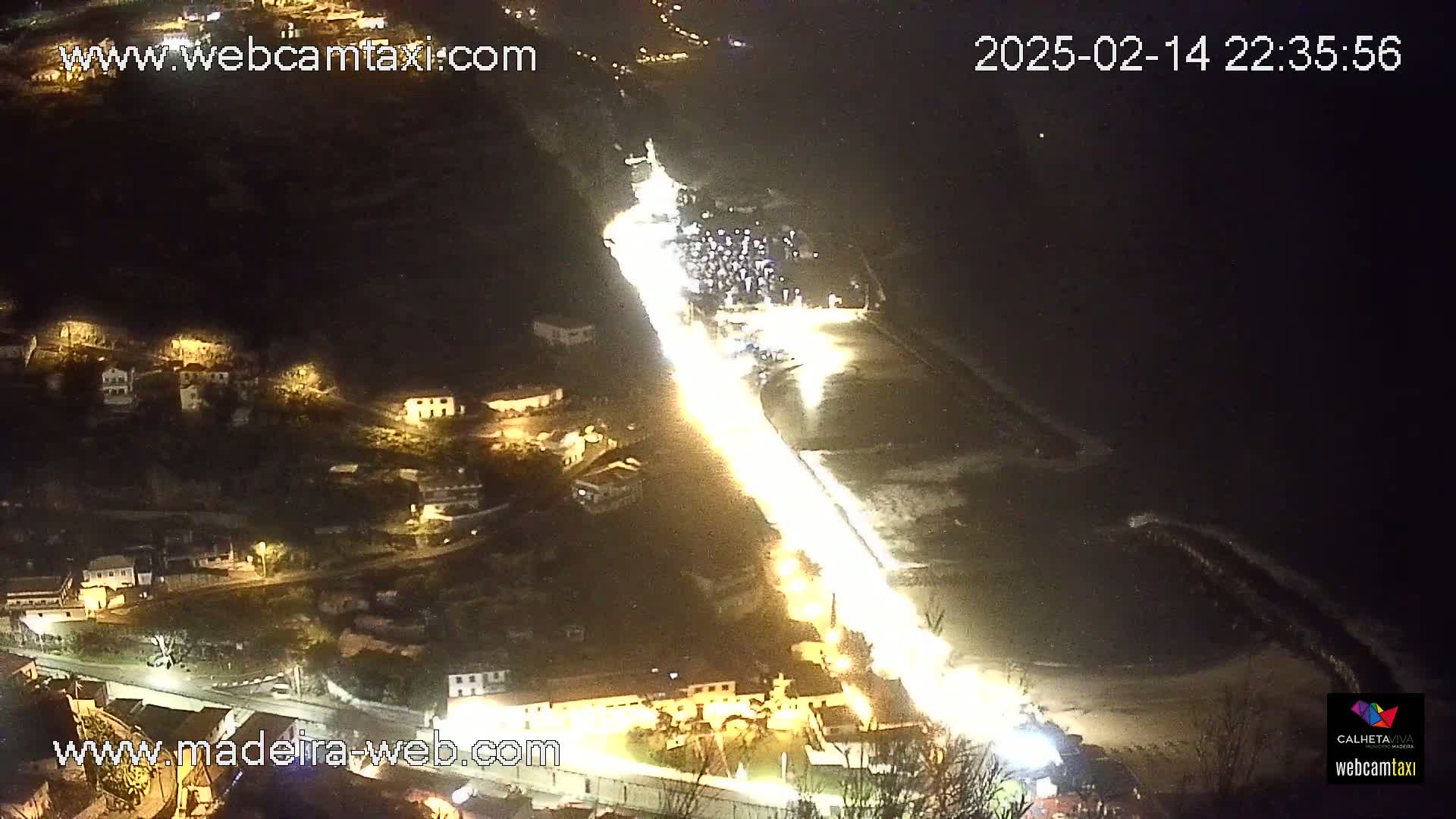 A brightly lit road winds down a hillside toward a dark, wavy ocean at night.