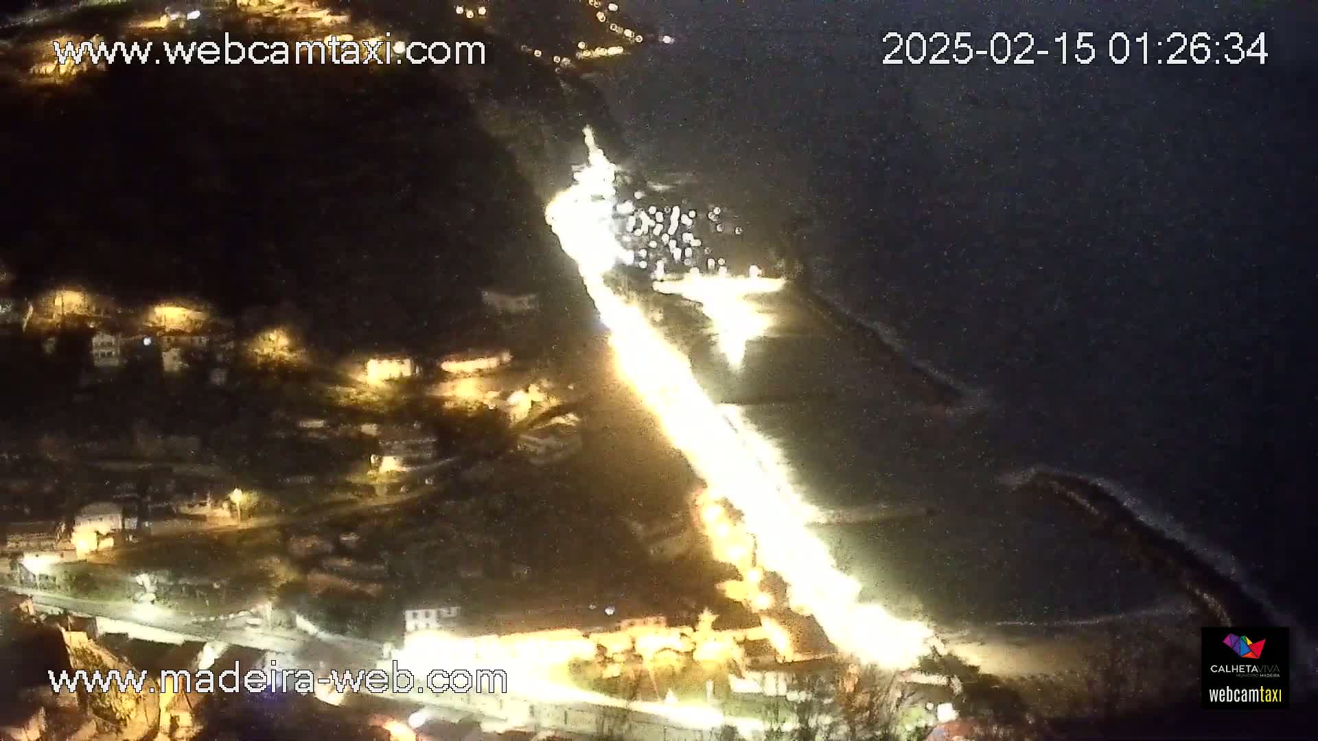 A nighttime aerial view shows a brightly lit road winding down a hillside toward a dark body of water with a small collection of boats.