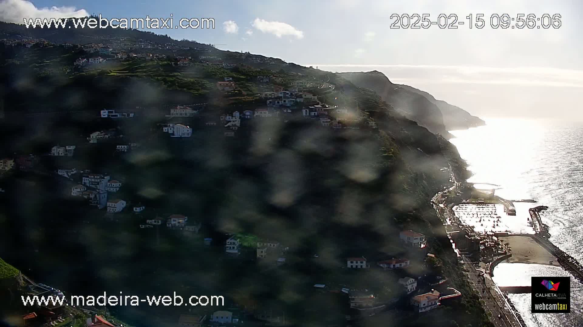 A coastal town nestled on a hillside overlooking a calm ocean, viewed through a rain-streaked window.