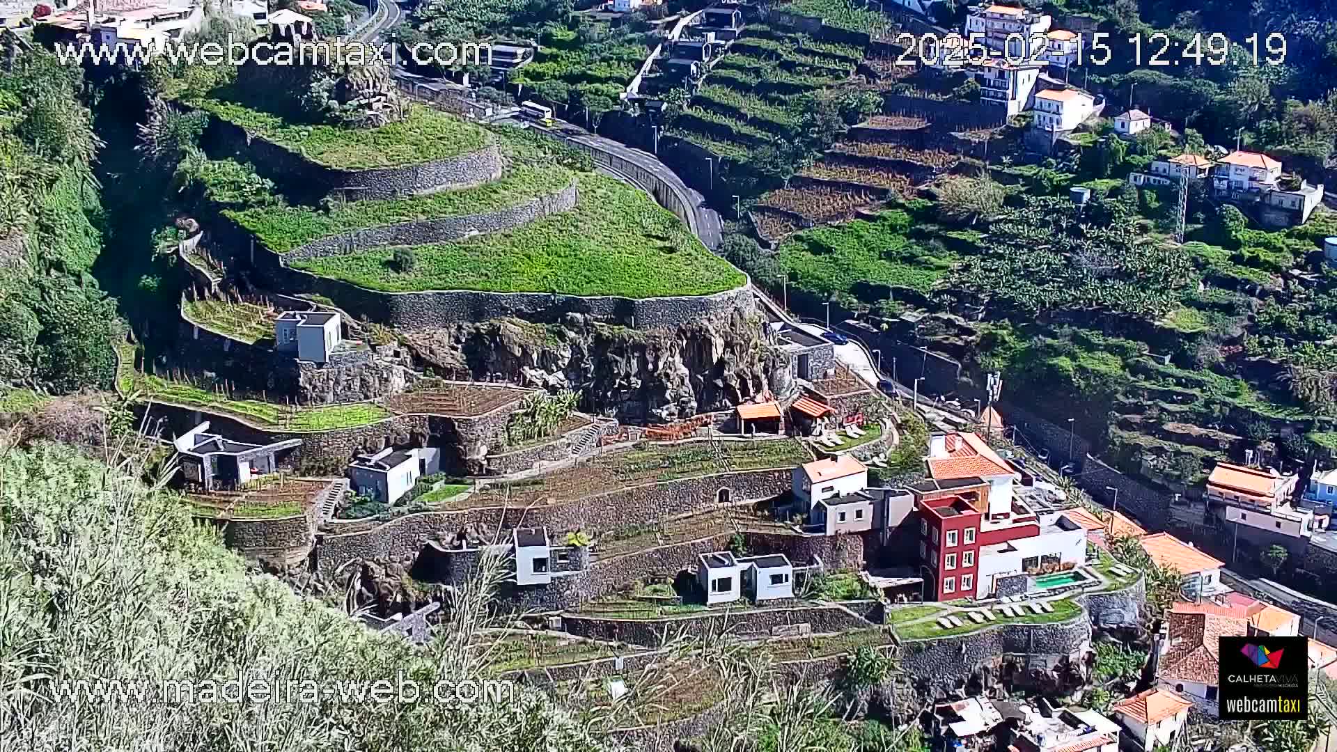 A hillside village with terraced gardens and homes is seen on a sunny day, with a road winding through the landscape.