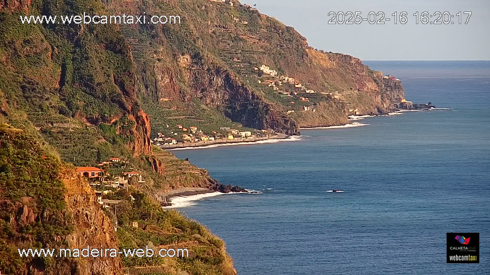 A coastal village clings to the side of a steep, verdant cliff overlooking a calm, blue ocean under a clear sky.