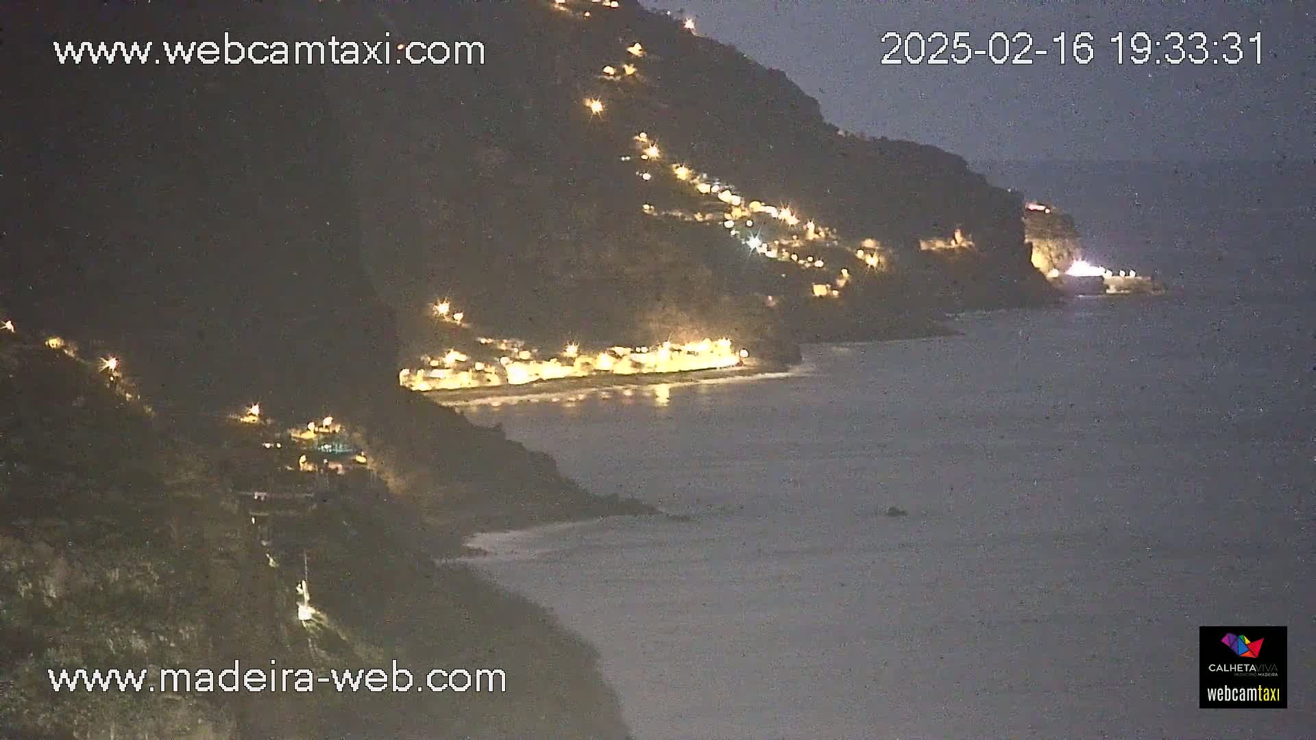A nighttime coastal scene shows a dark hillside dotted with lights descending to a beach and calm ocean.