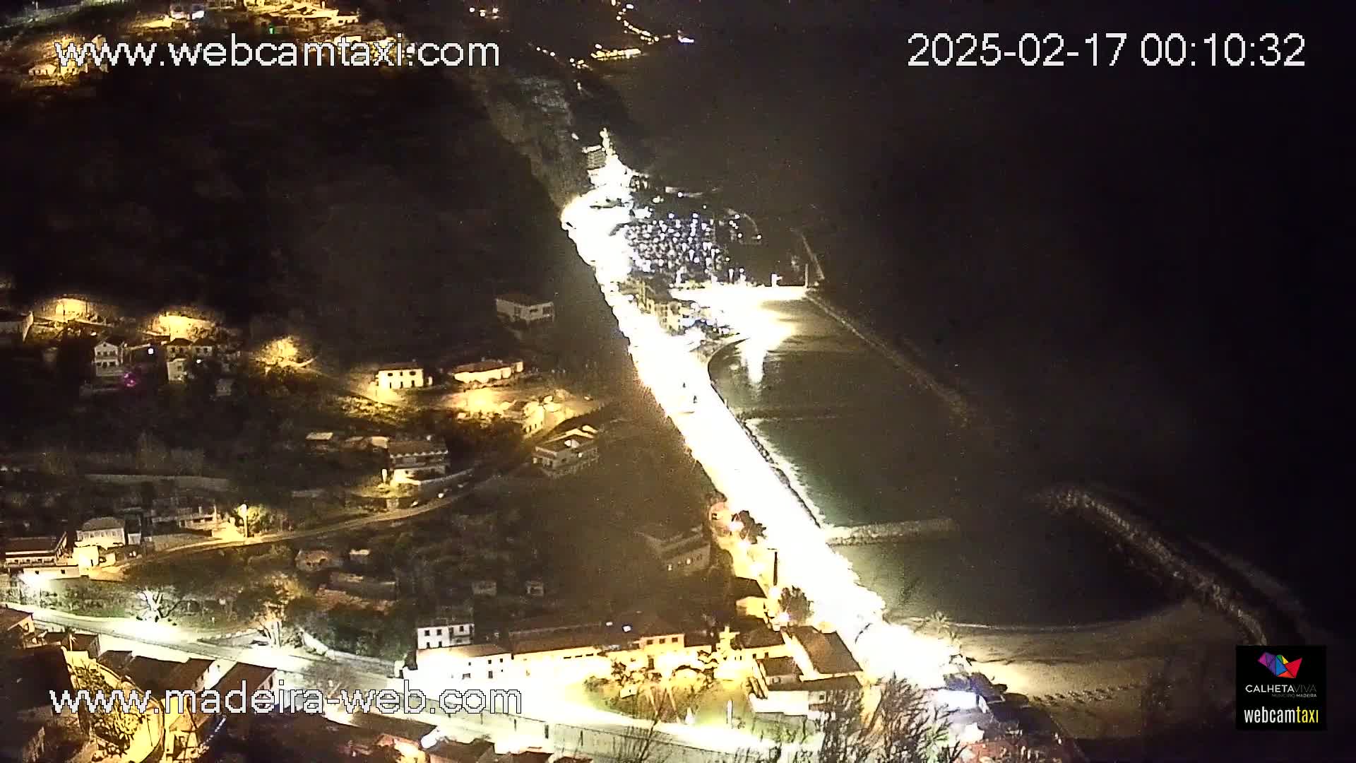 A nighttime, high-angle view shows a coastal town illuminated with lights, extending along a dark, gently curving shoreline next to a calm body of water.