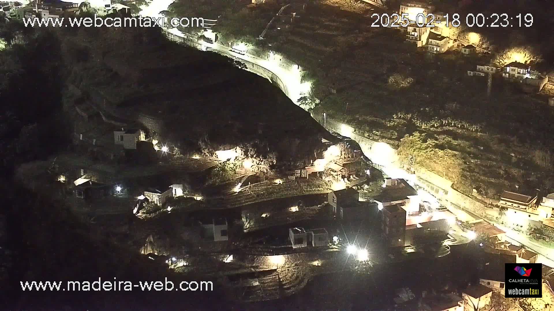An aerial nighttime view shows a cluster of buildings nestled into a hillside, with a road winding through the landscape.