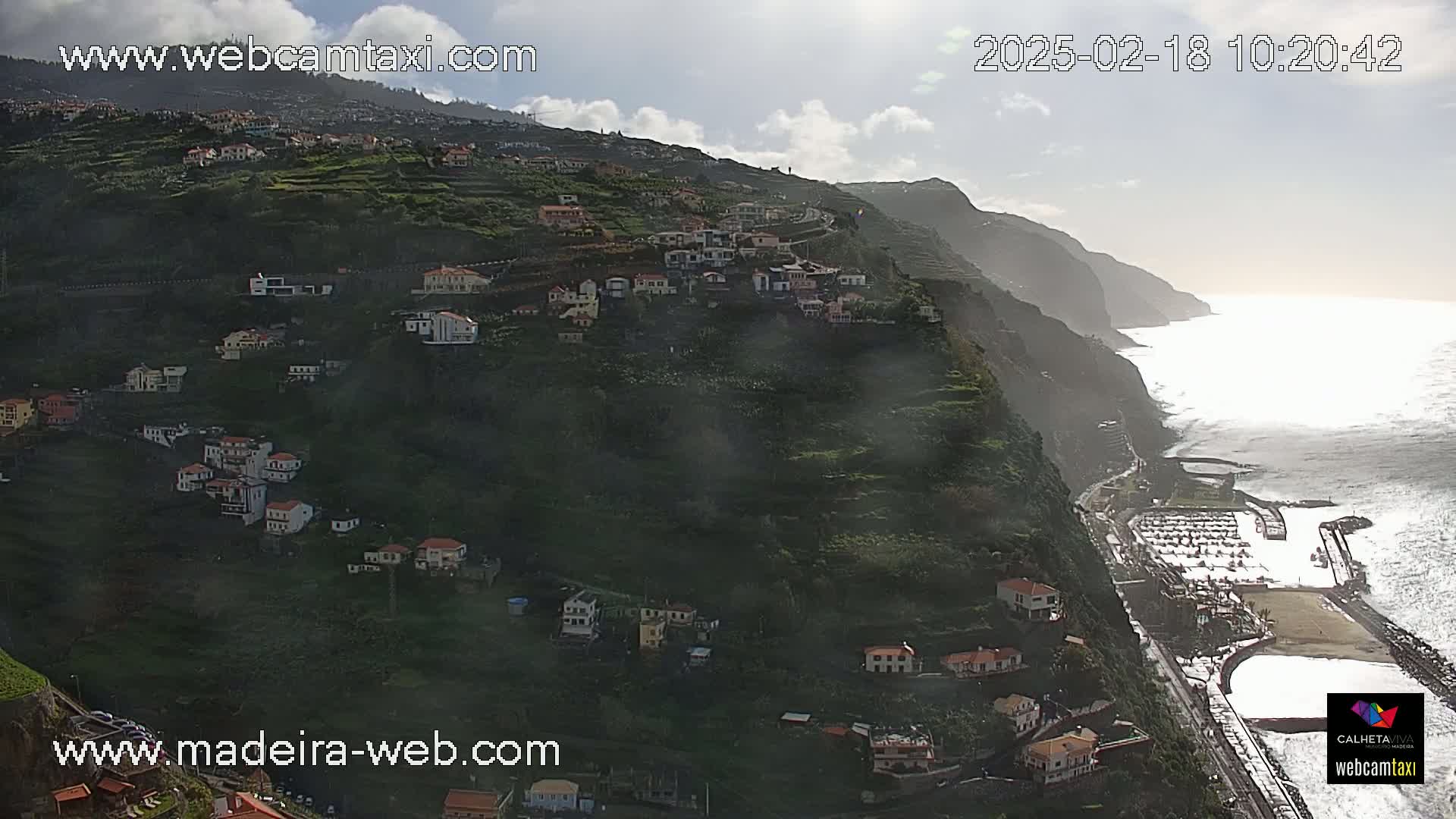A hillside town clings to a steep, verdant cliff overlooking a calm ocean under a partly cloudy sky.