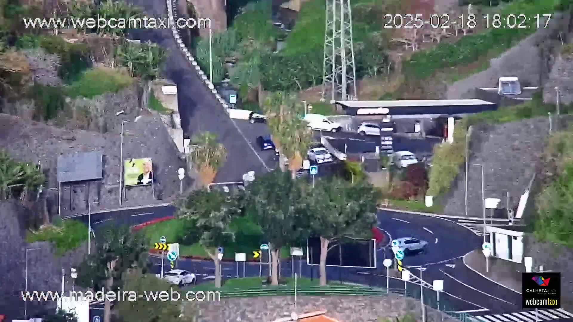 An aerial view shows a curvy road with several cars and a gas station nestled into a hillside on a partly cloudy day.
