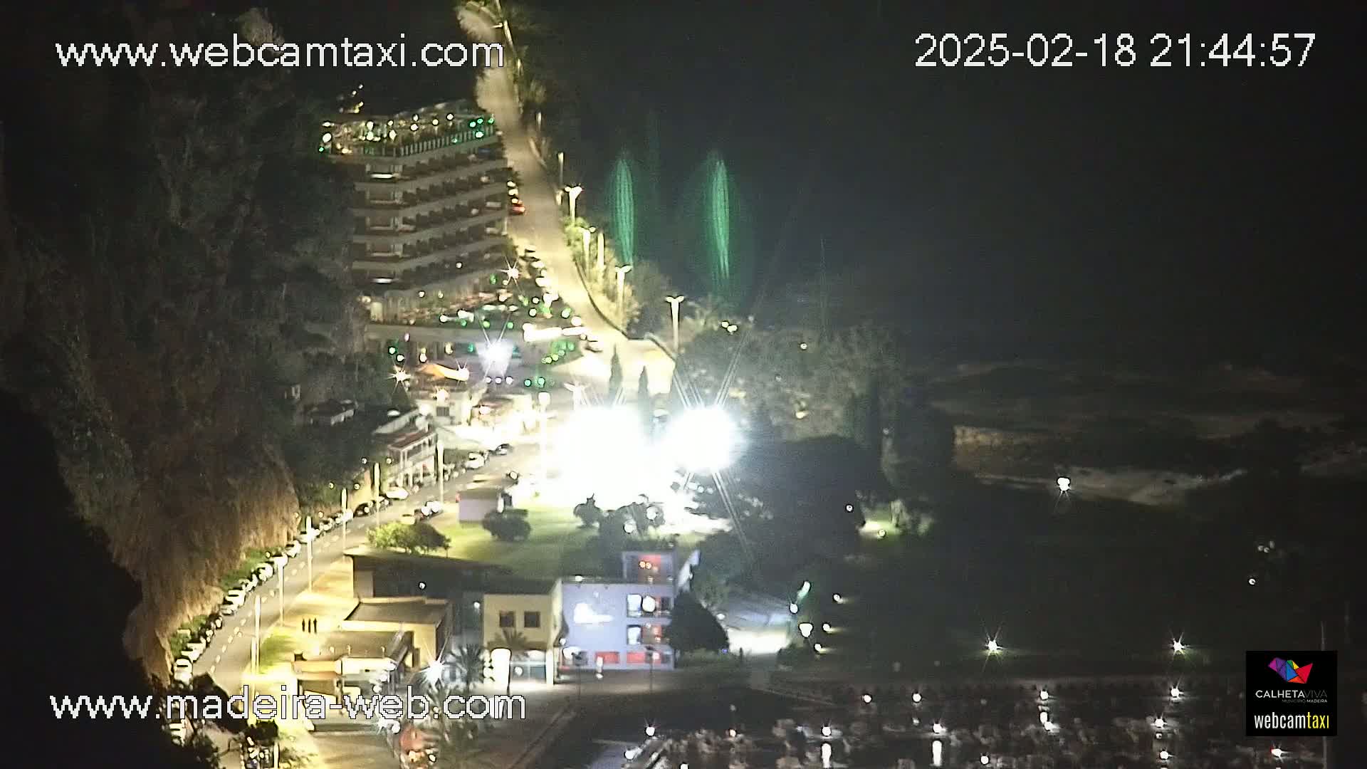 A nighttime aerial view of a coastal town shows a parking lot, buildings, a roadway, and a marina, with two green fountains visible near the water.