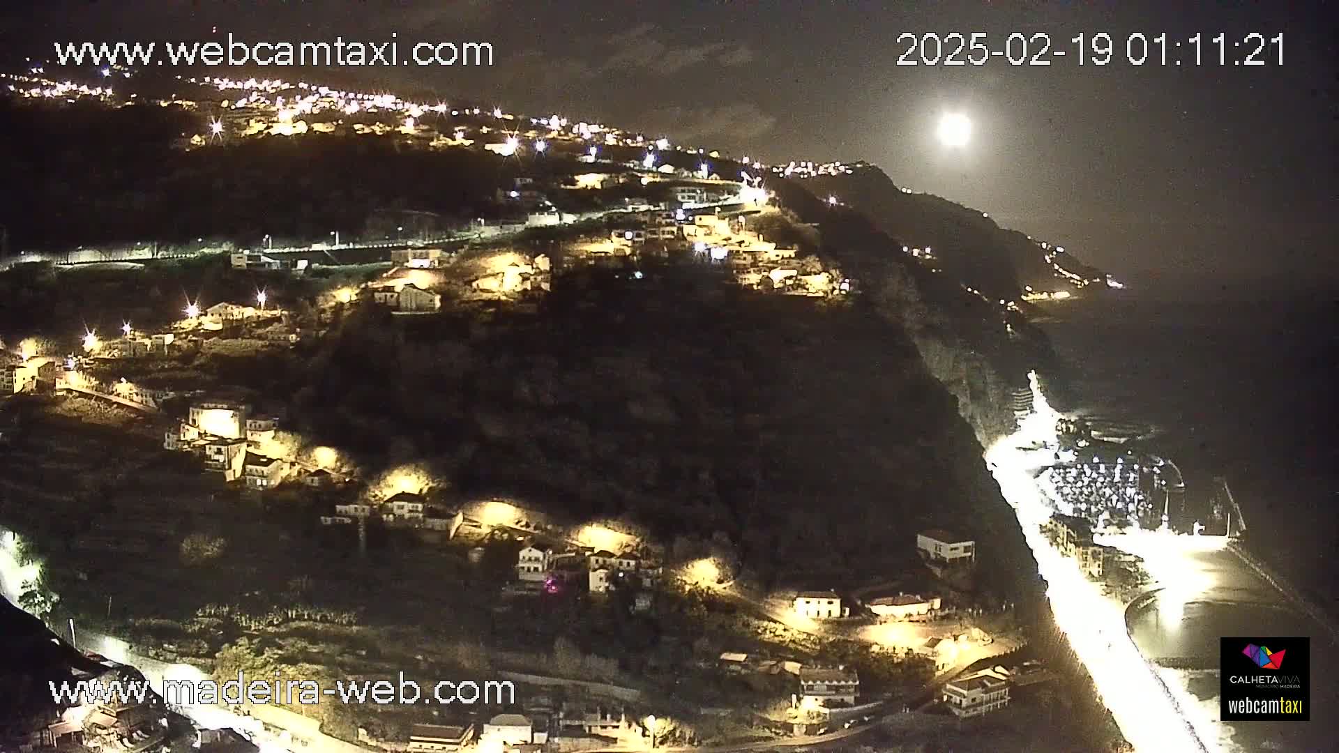 A nighttime, high-angle view shows a hillside town illuminated by streetlights, extending down to a bright coastal road and a dark ocean under a bright moon.