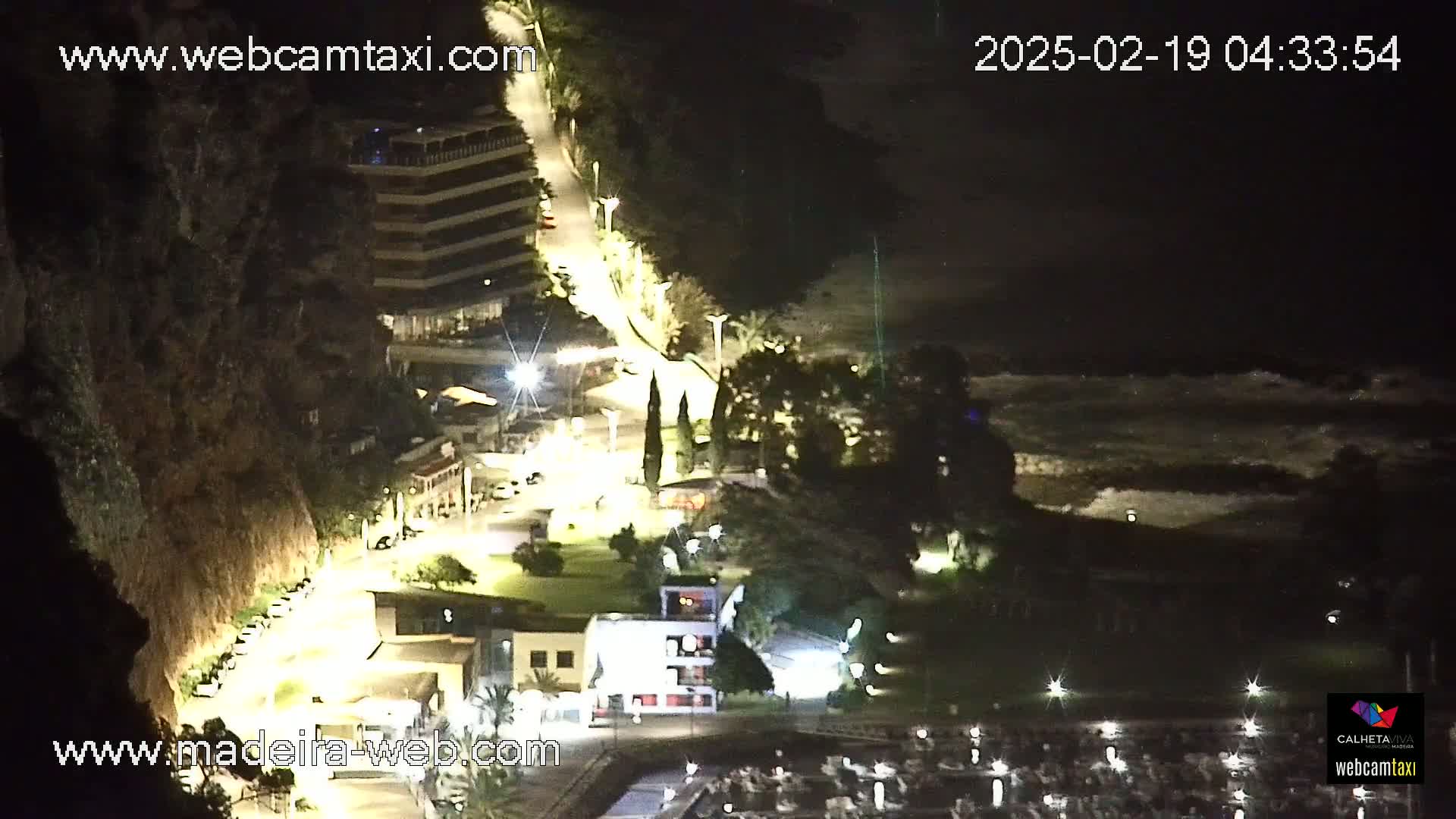 A nighttime coastal scene shows a lit roadway winding down a cliffside toward a marina, with dark, wavy water visible beyond.