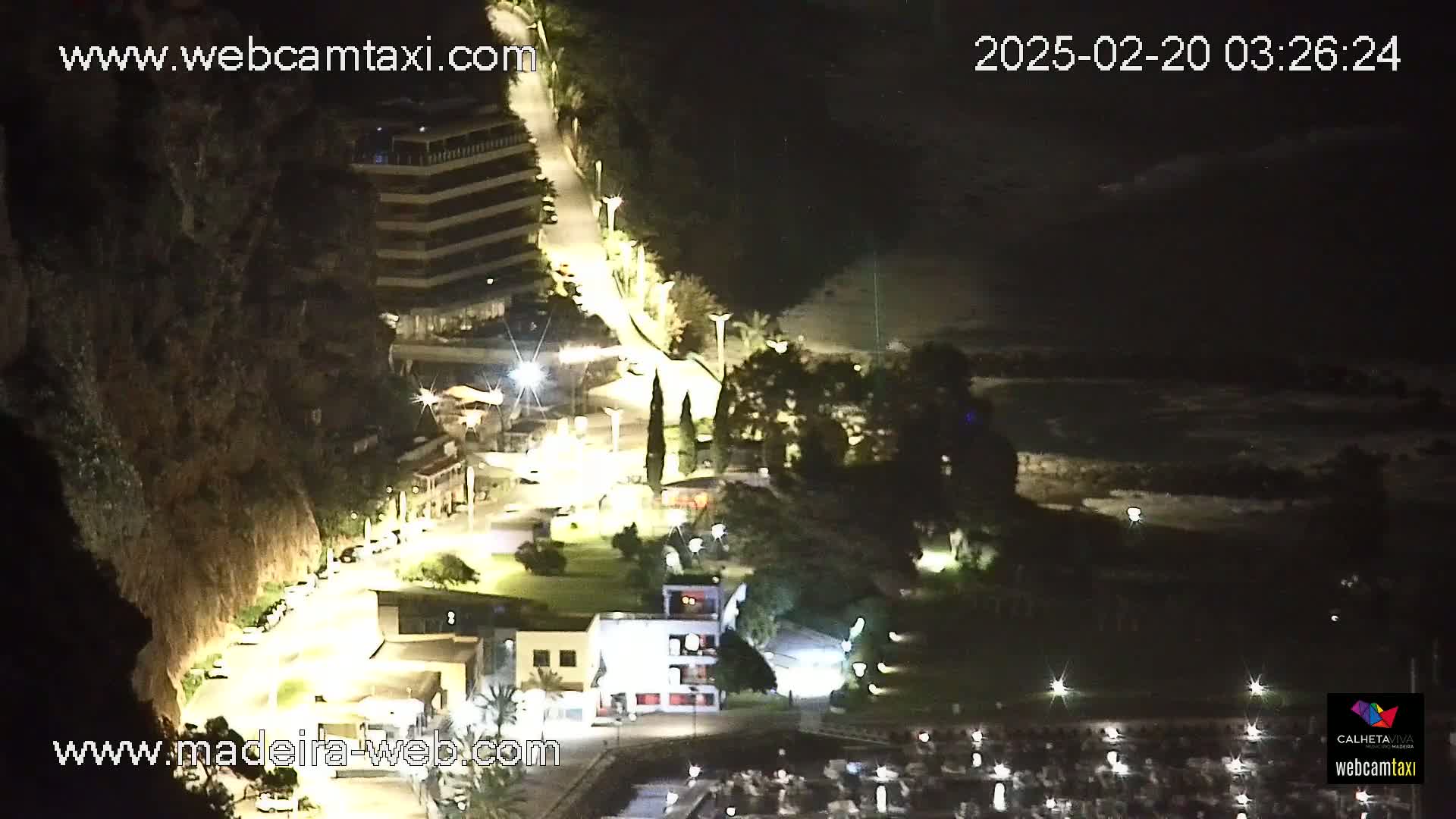 A nighttime, coastal scene shows a winding road lit by streetlights leading down to a marina, with a multi-story building perched on a cliffside.