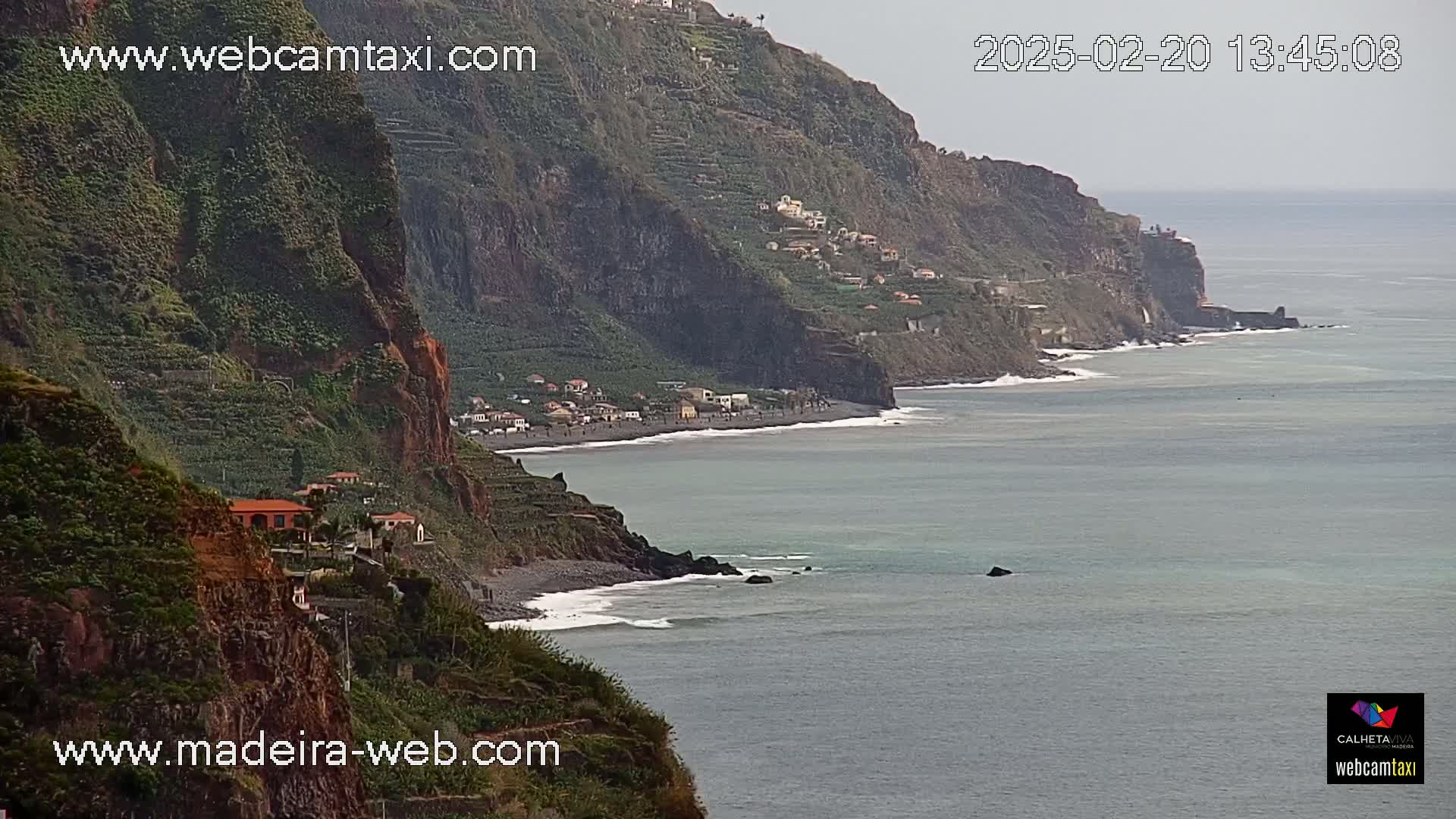 A coastal view shows a dark, rocky cliffside with sparse vegetation and a few buildings overlooking a calm ocean with small waves lapping the shore.