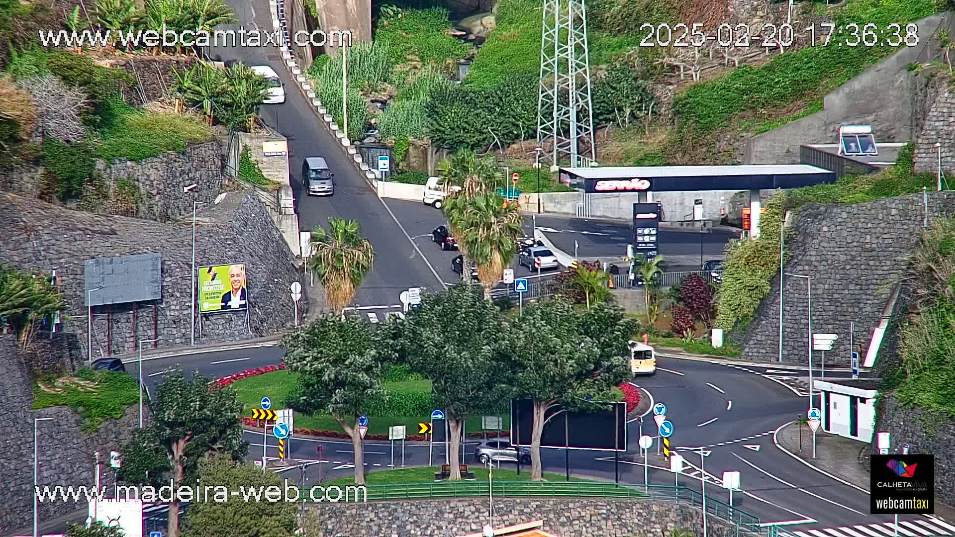 A sunny, high-angle view of a roundabout and intersecting roads with several cars and a gas station amidst lush green landscaping and stone walls.