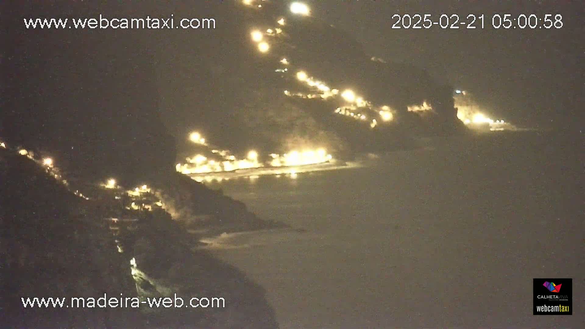 A nighttime coastal scene shows a dark, hilly coastline with scattered lights along the shore and on the hillsides, overlooking a dark body of water.