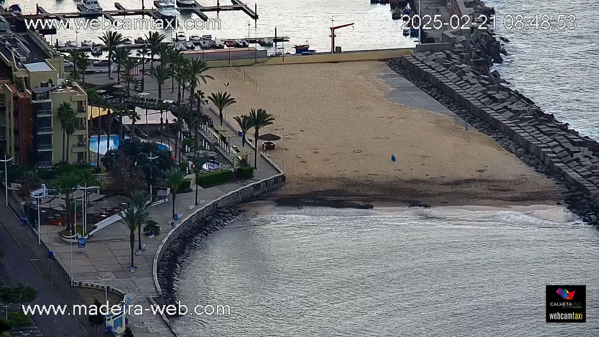 An aerial view shows a sandy beach next to a calm ocean, bordered by a stone wall and a building with a swimming pool, surrounded by palm trees.