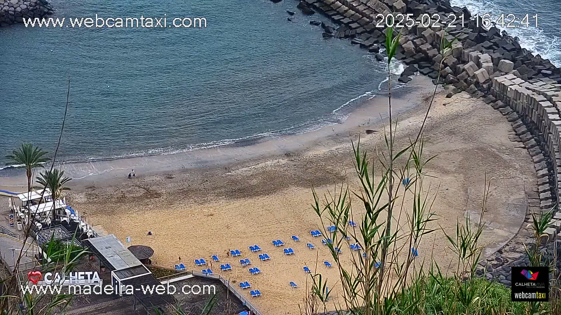 An aerial view shows a sandy beach with blue lounge chairs and calm ocean waves lapping the shore, partially sheltered by a rock wall.