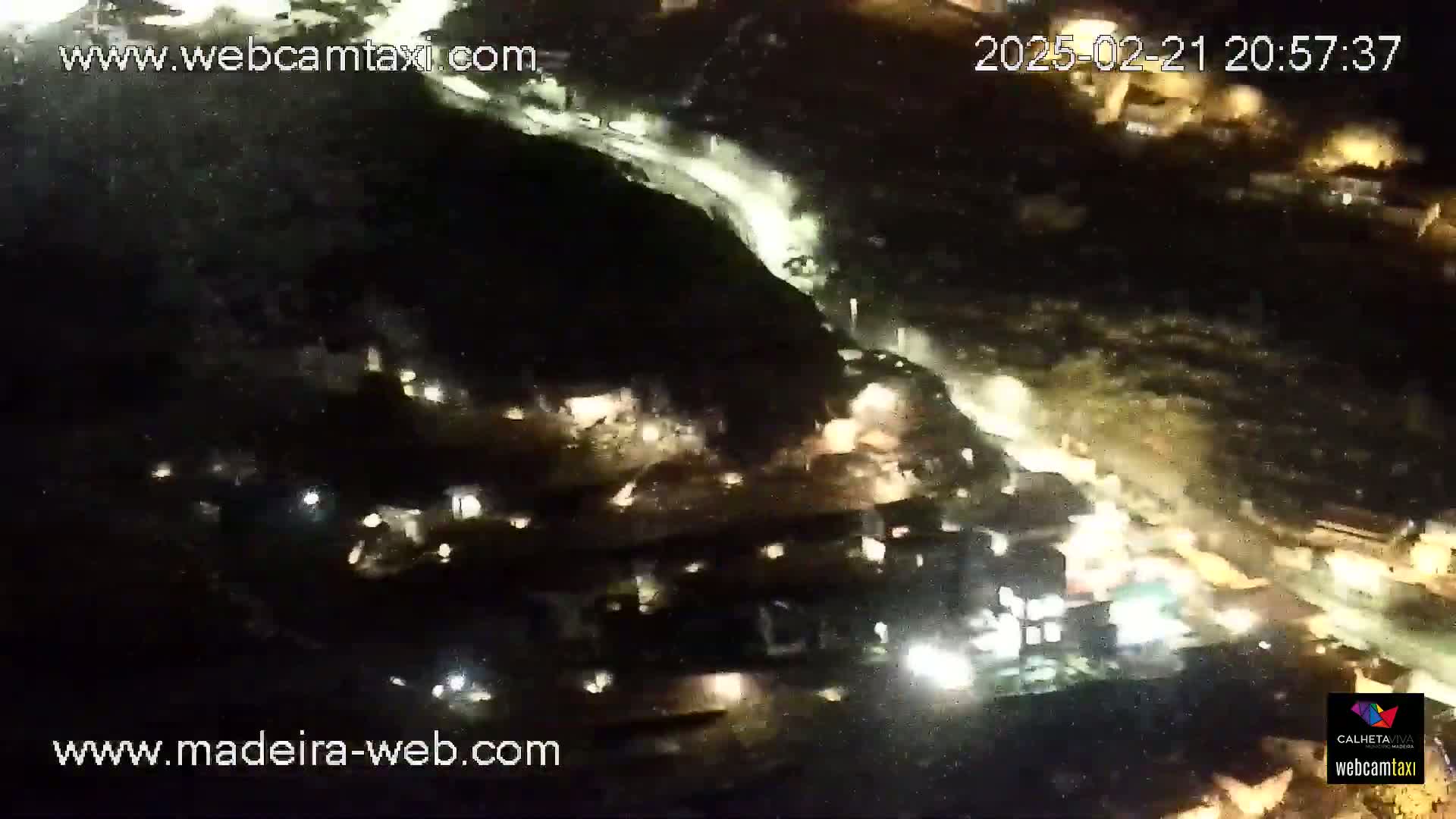 A nighttime aerial view shows a hilly area with sparsely lit buildings and a winding road with vehicle lights.