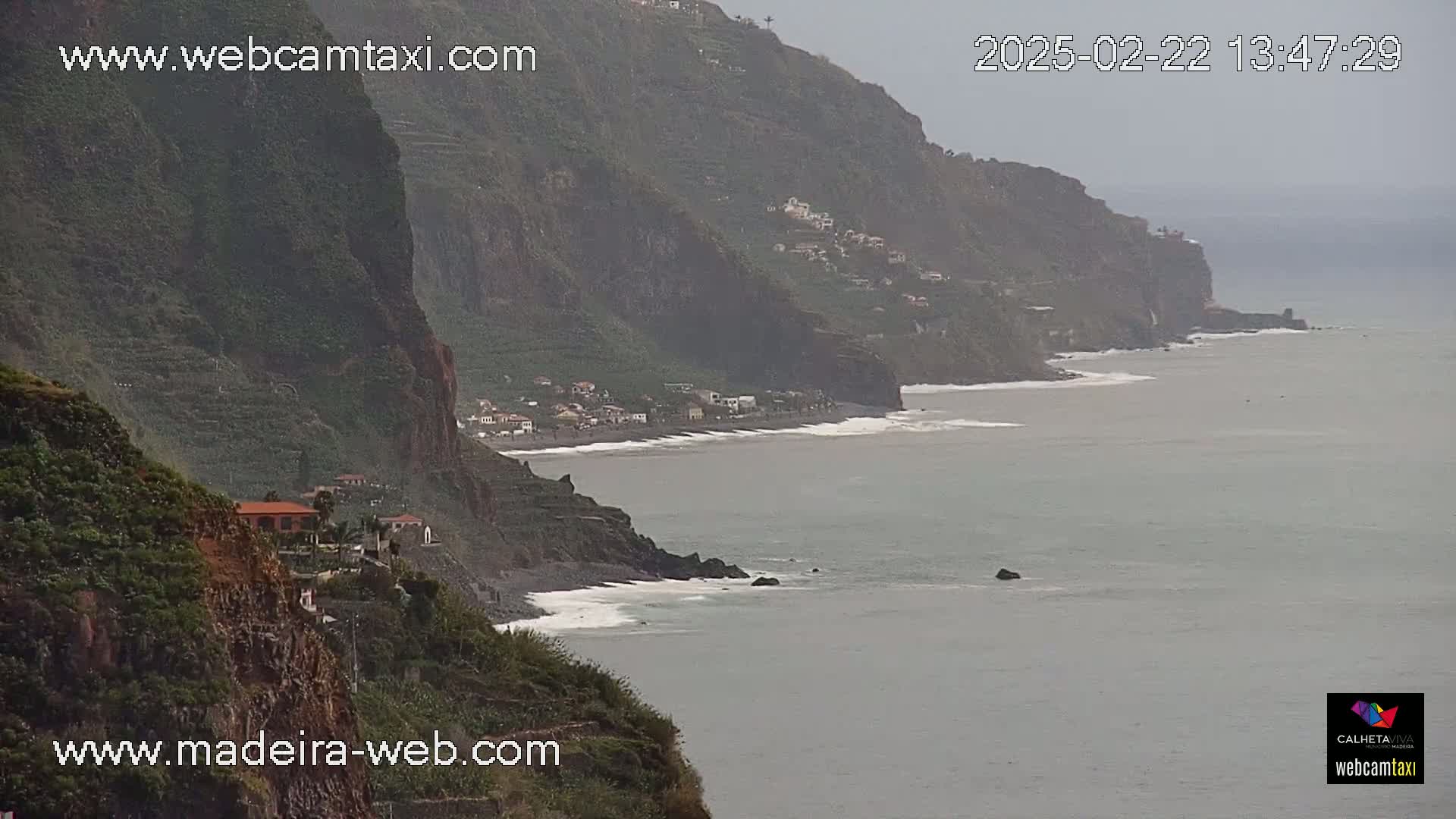 A coastal view shows a rocky cliffside with a few buildings overlooking a beach and ocean, under an overcast sky.