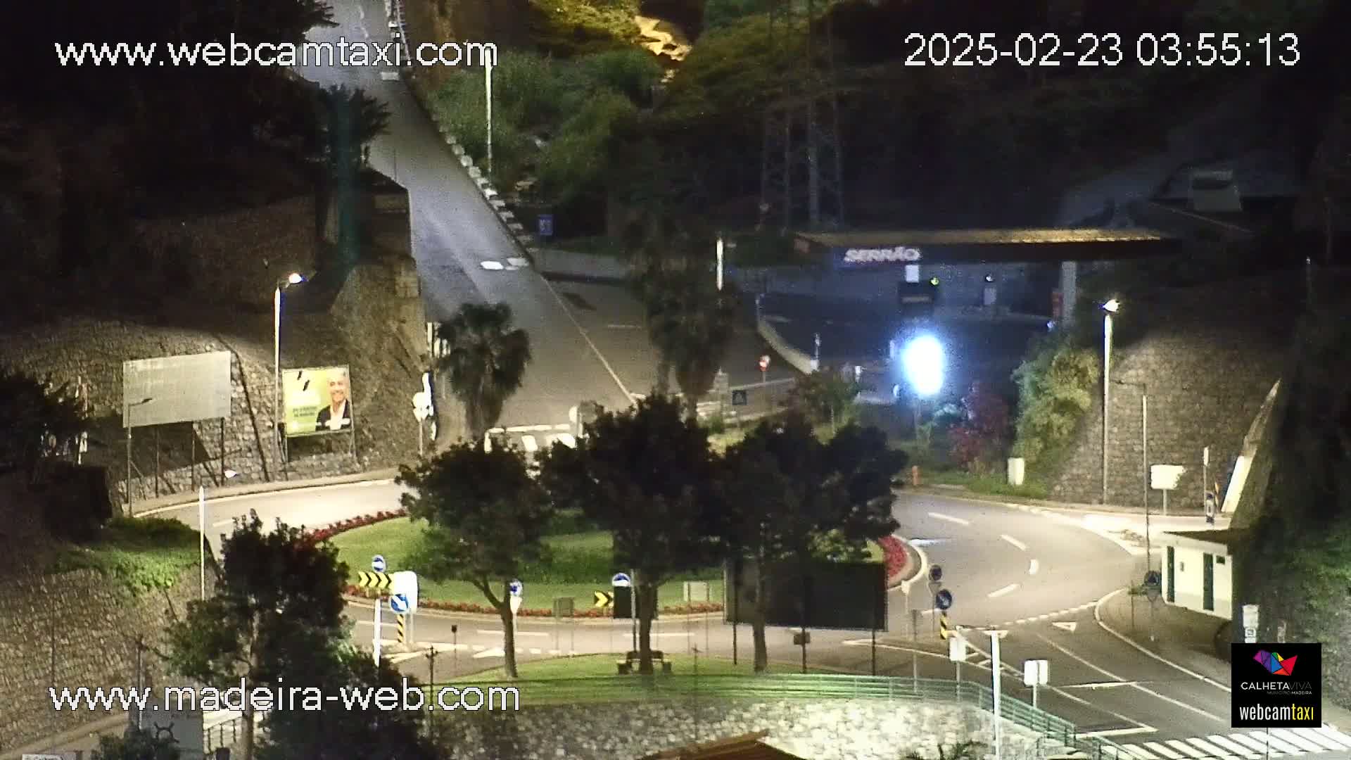 A nighttime, overhead view of a roundabout and roadway intersection, with surrounding vegetation and buildings illuminated by artificial light.