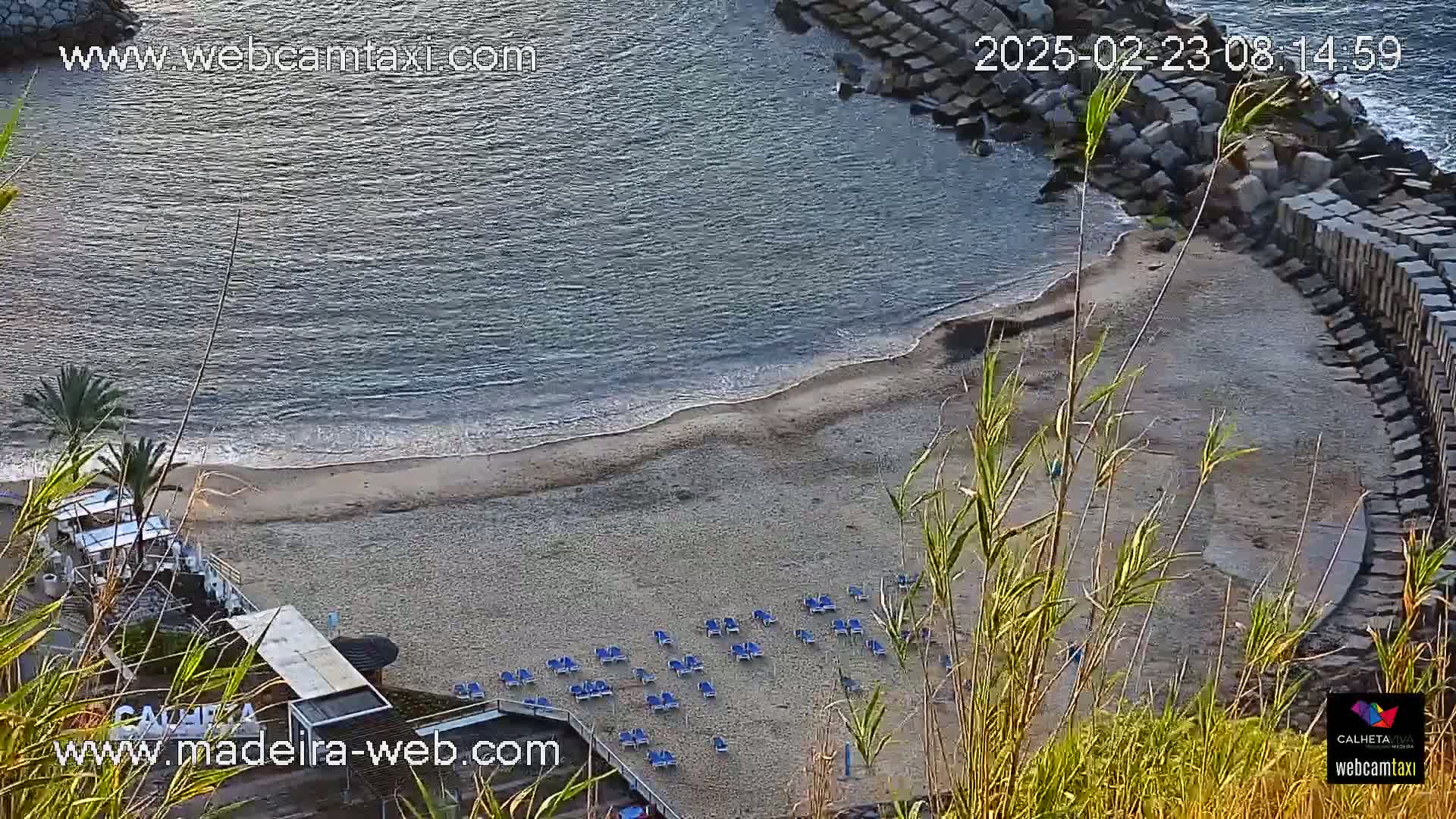An aerial view shows a sandy beach with calm ocean waves and numerous empty lounge chairs under a clear sky.