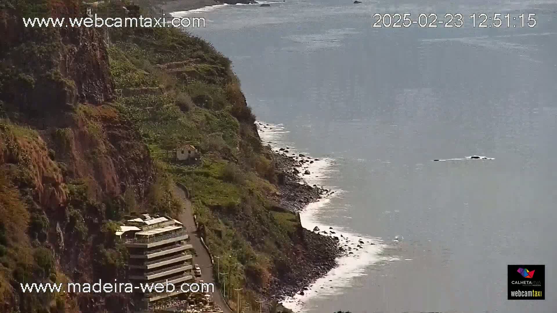 A rocky, green cliffside with a road and buildings overlooks a calm ocean with small waves breaking on a dark, pebbly shore.
