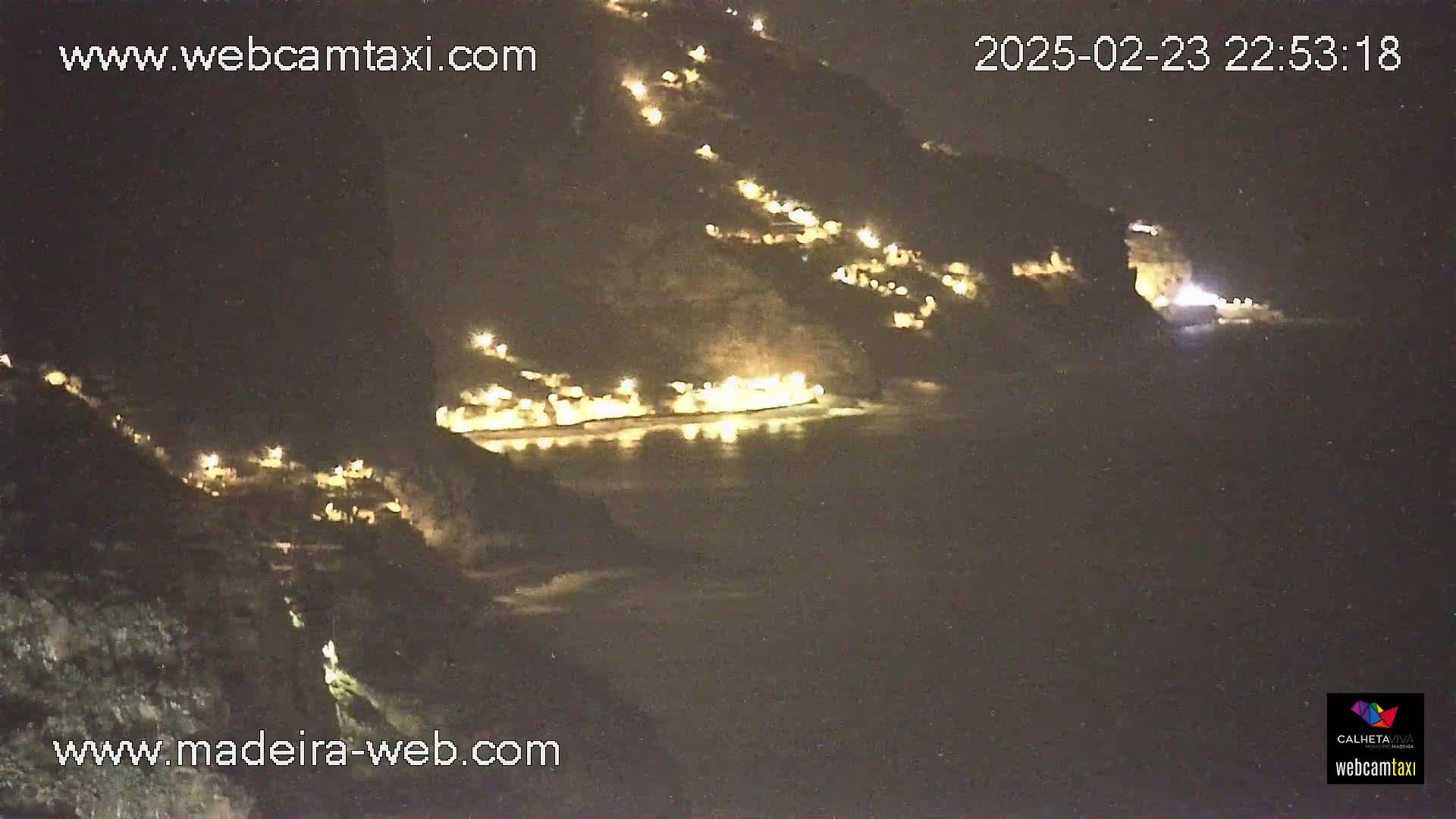 A nighttime view of a coastal town illuminated by lights, nestled against a dark hillside and overlooking a dark body of water.