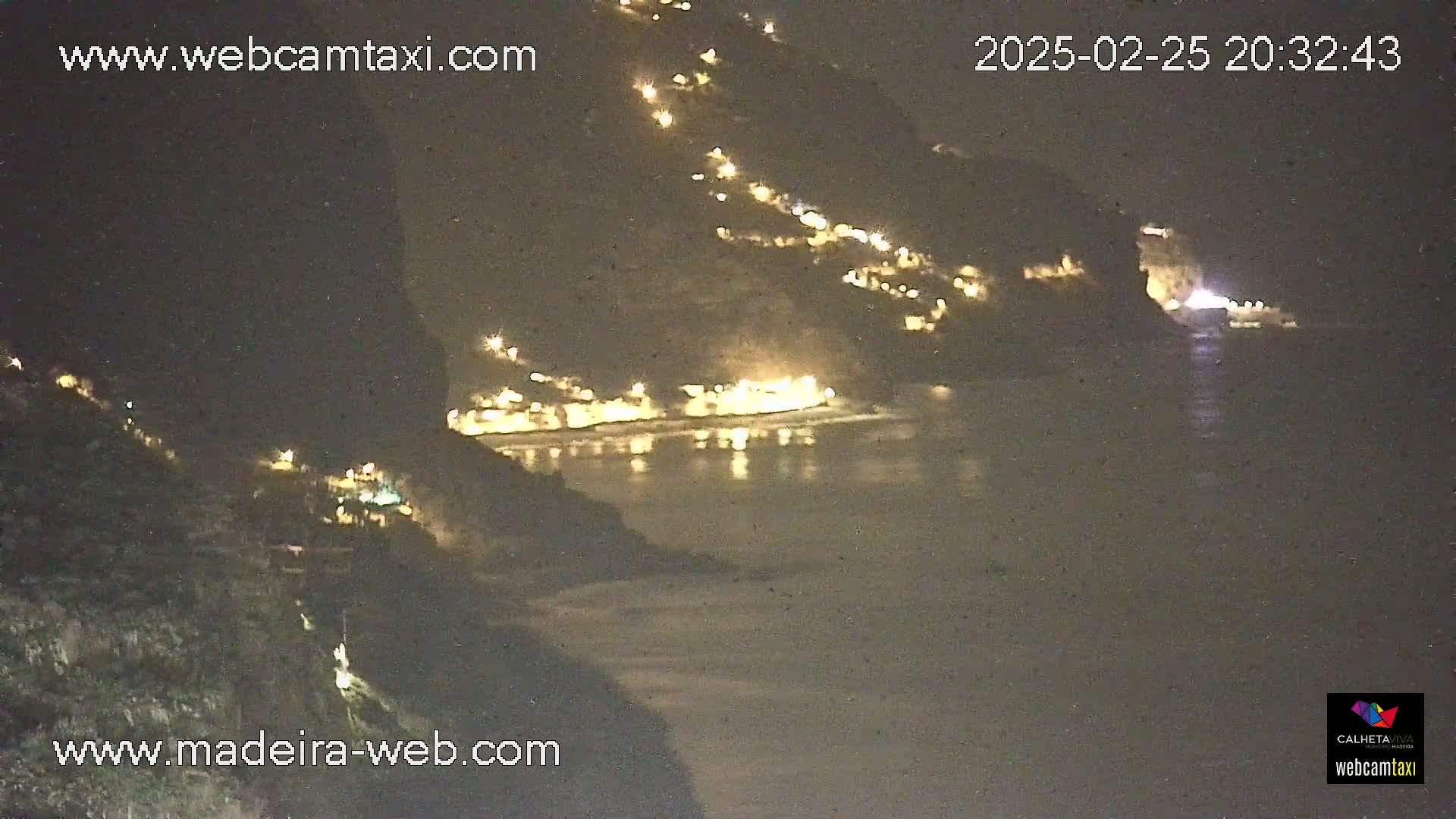 A nighttime view of a coastal town nestled between dark cliffs, with its lights illuminating the water below.