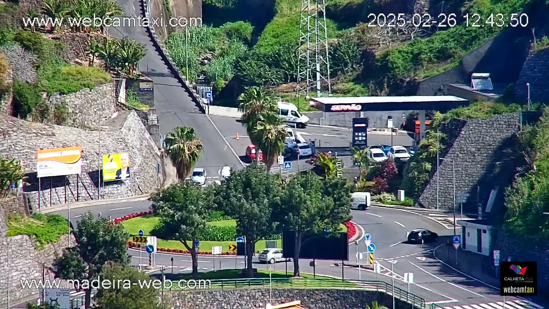 A sunny day overlooking a roadway with several cars and a gas station, surrounded by stone walls and palm trees.