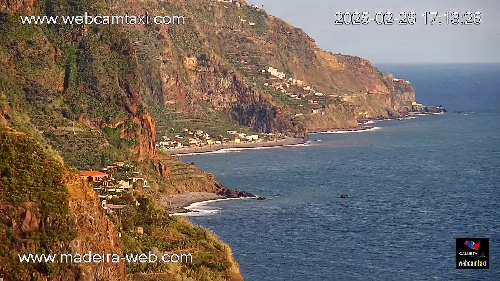 A rocky, vegetated coastline with a small village and a dark-sand beach meets a calm ocean under a clear sky.