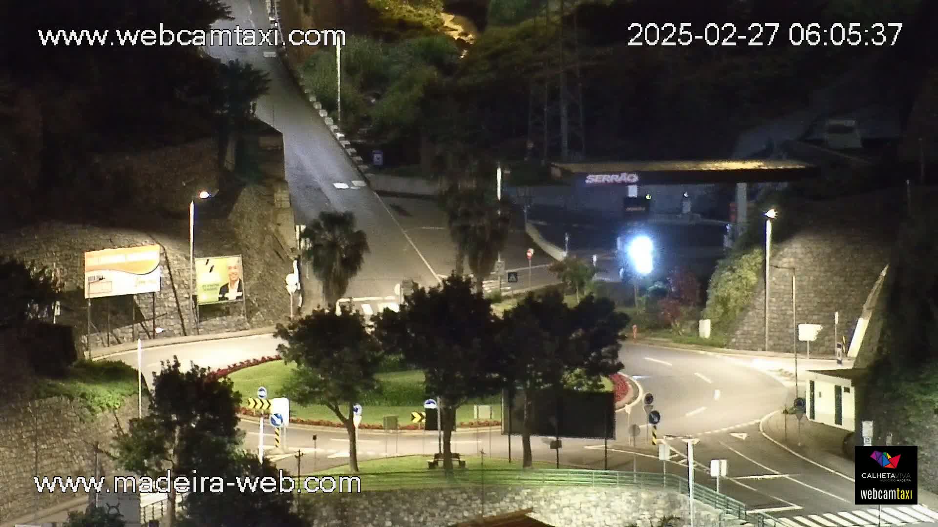 A nighttime view of a roundabout and curving road illuminated by streetlights, nestled among trees and stone walls.