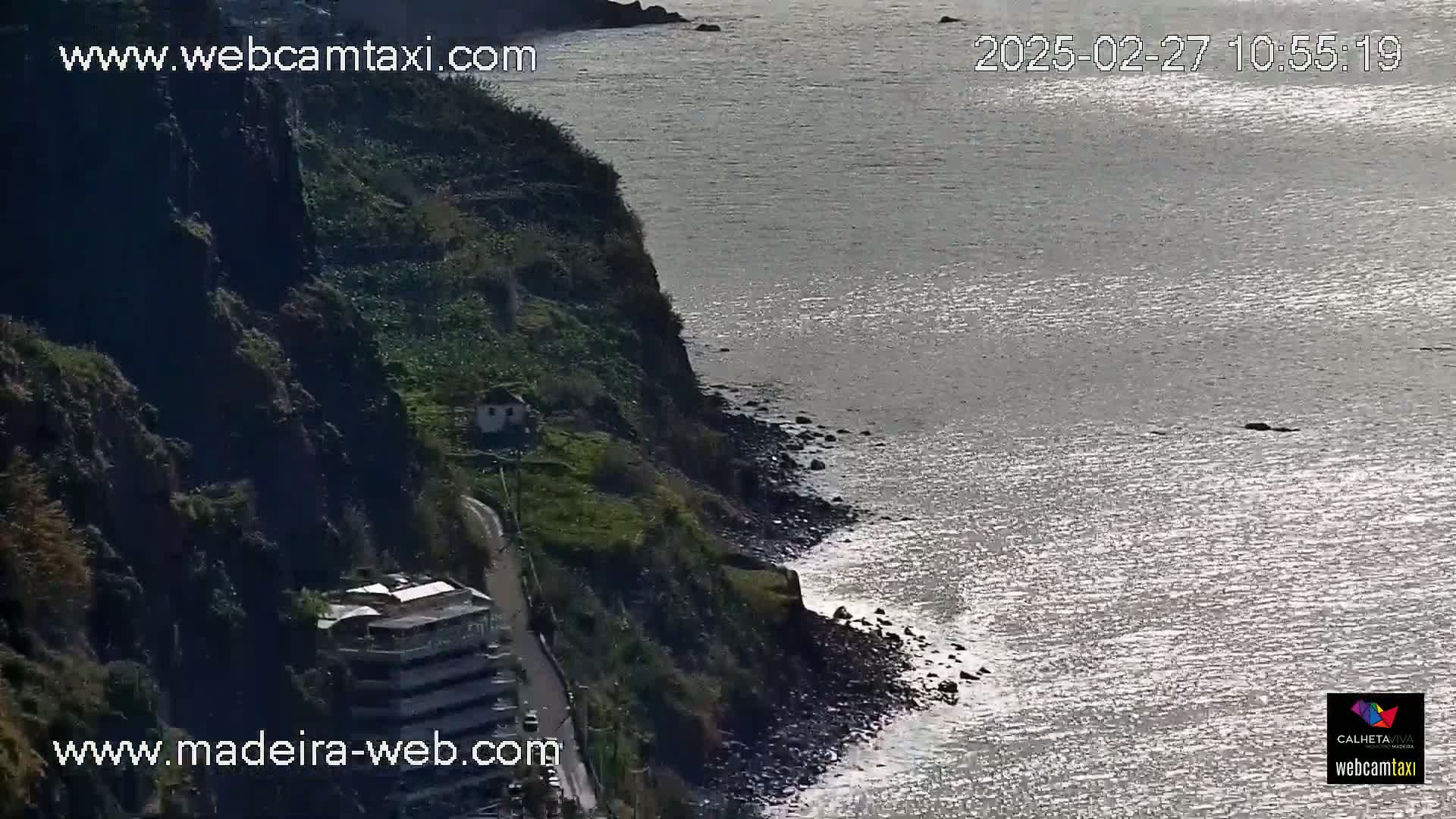 A rocky cliffside with a road and buildings overlooks a calm, sunlit ocean.