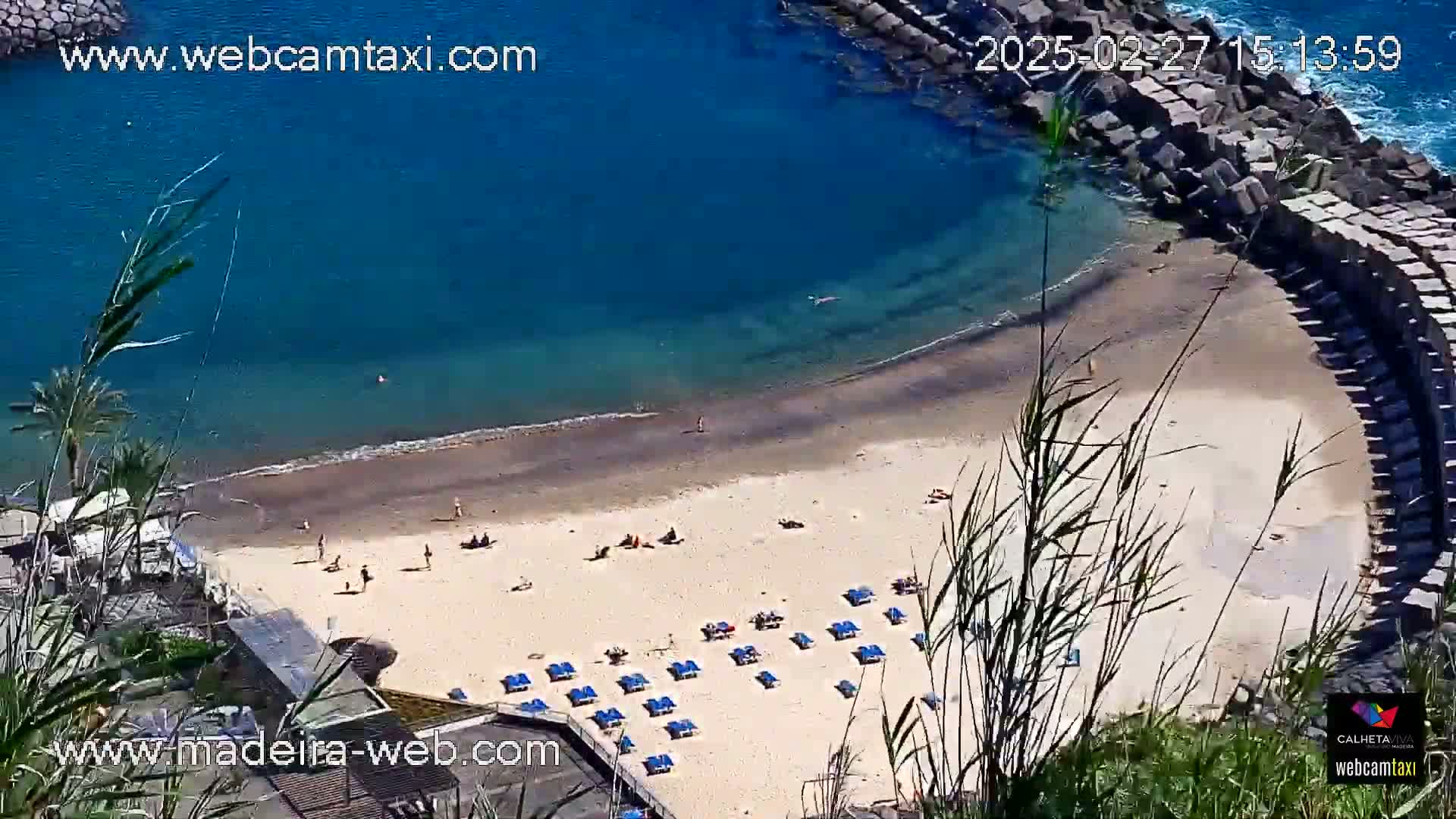 An aerial view shows a sandy beach with scattered sunbathers and lounge chairs, partially enclosed by a rock wall, and bordered by calm, clear blue ocean water.