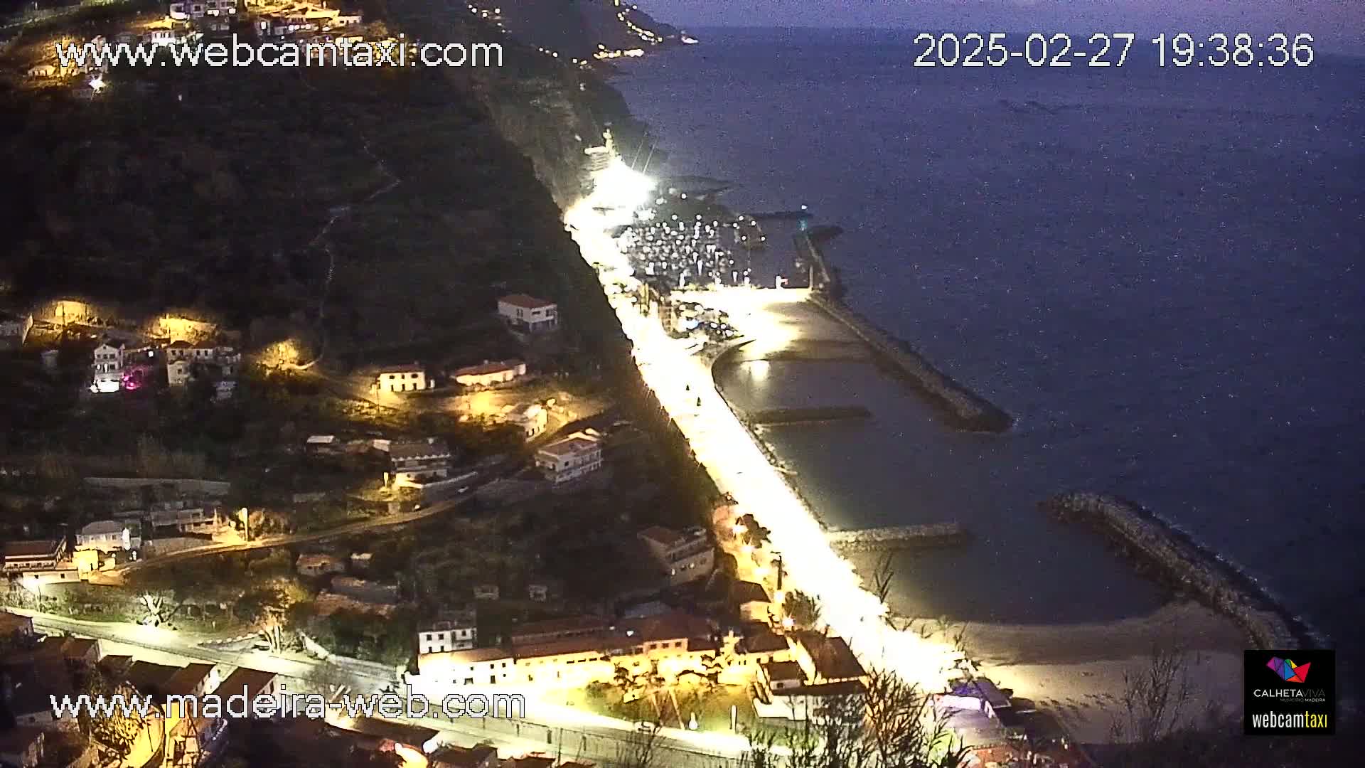 A nighttime coastal village scene shows a brightly lit roadway curving along a shoreline next to a calm, dark ocean, with buildings illuminated on a hillside above.