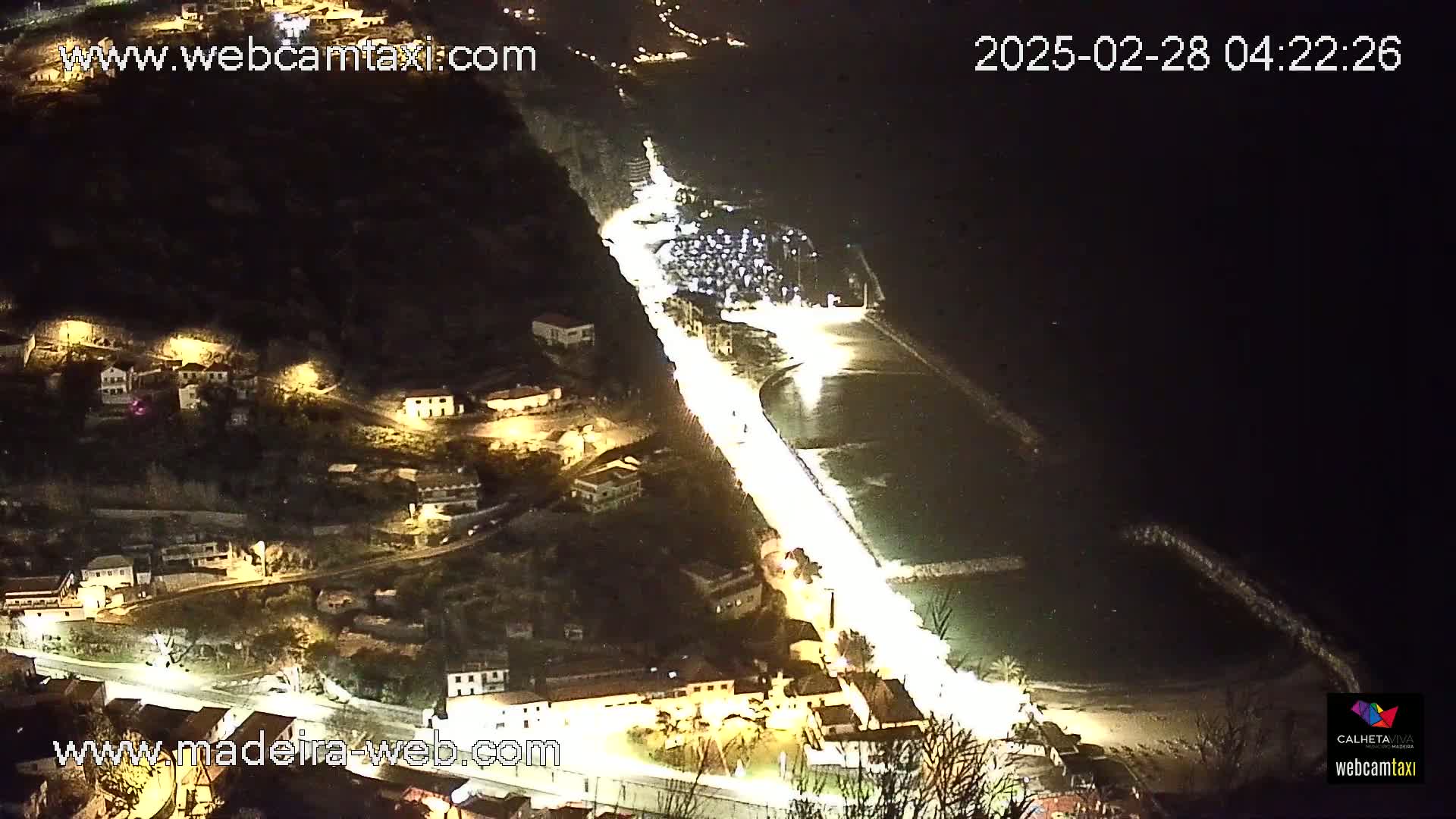 A nighttime, high-angle view shows a coastal town with illuminated streets and buildings along a dark ocean.