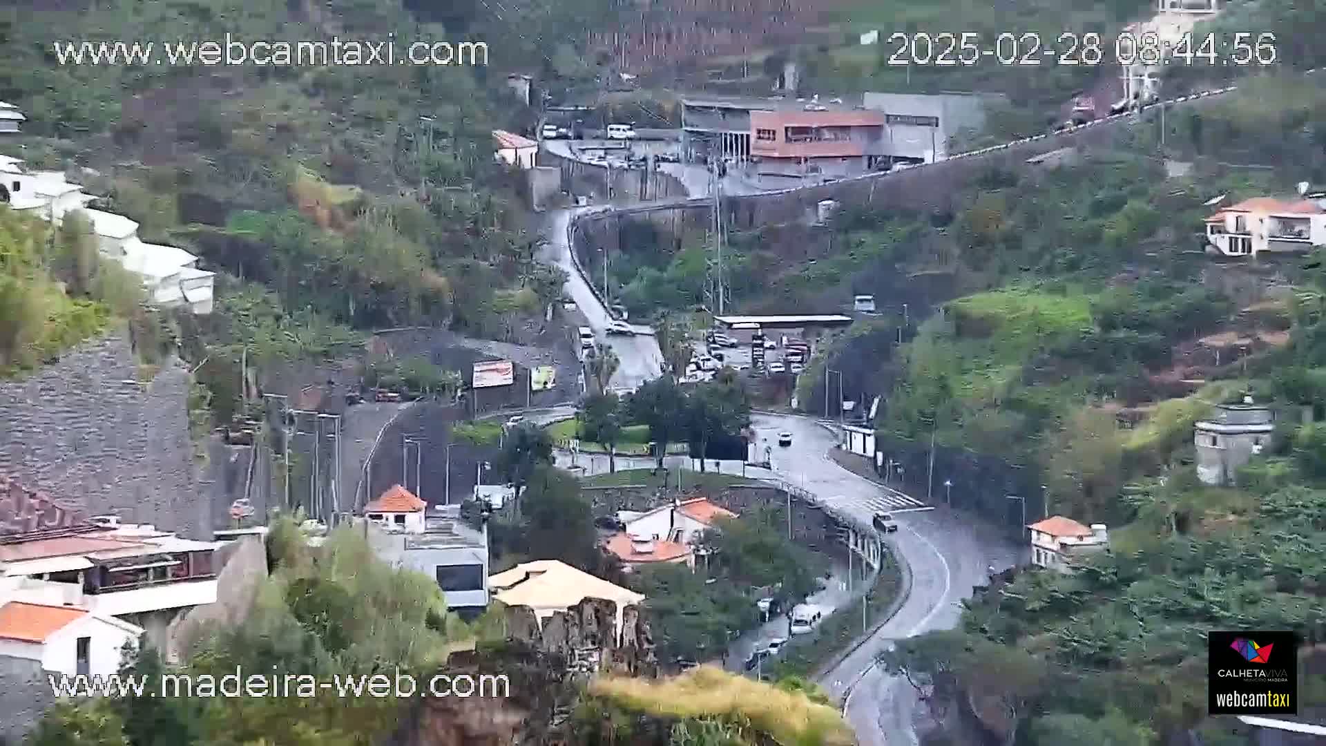 A winding road through a mountainous area with buildings and some sparse vegetation, under overcast conditions.