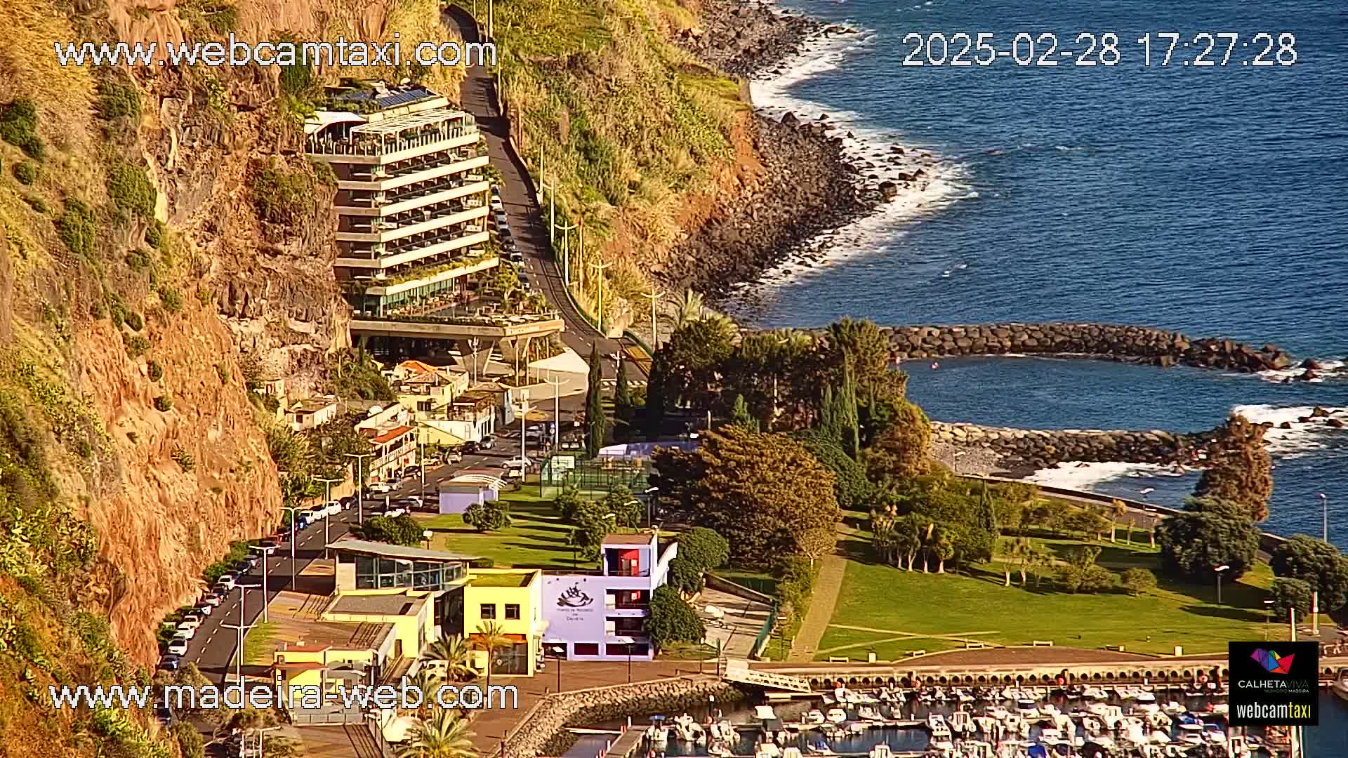 A coastal scene shows a multi-story hotel built into a cliff overlooking a calm ocean with a small harbor filled with boats and a park with green grass.