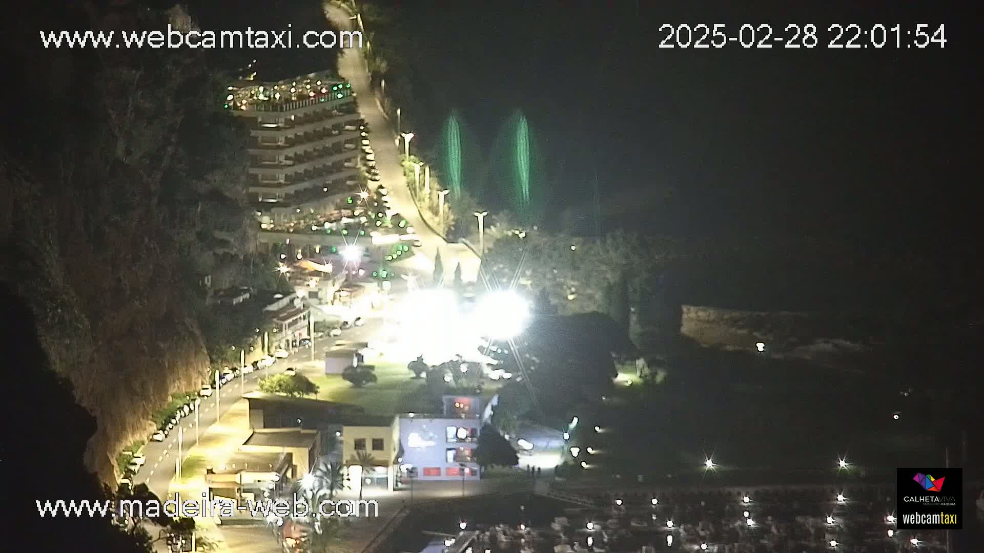 A nighttime aerial view of a coastal town shows a multi-story building, a road with parked cars, and a small harbor, with two vertical green lights visible in the distance.