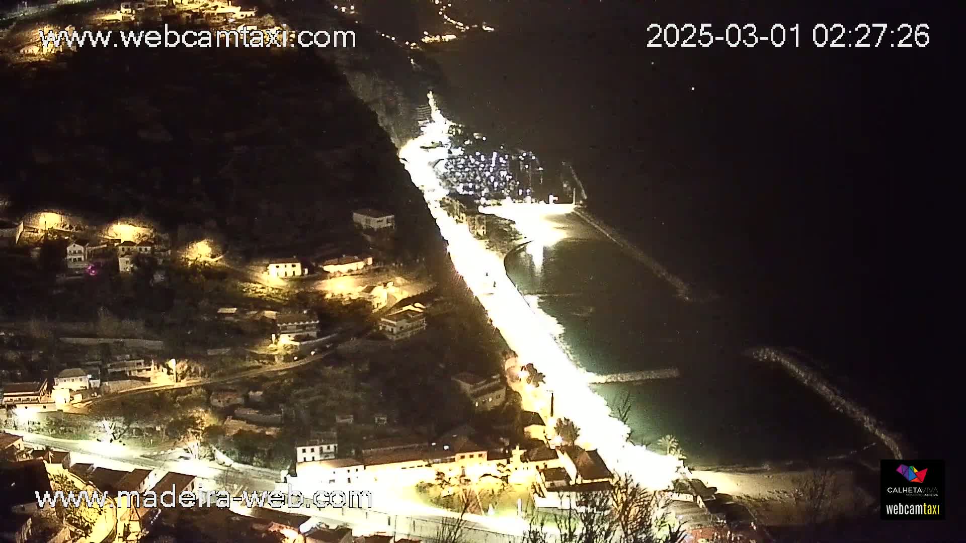 A brightly lit coastal road winds down a hillside at night, alongside a dark body of water with a beach and breakwater.